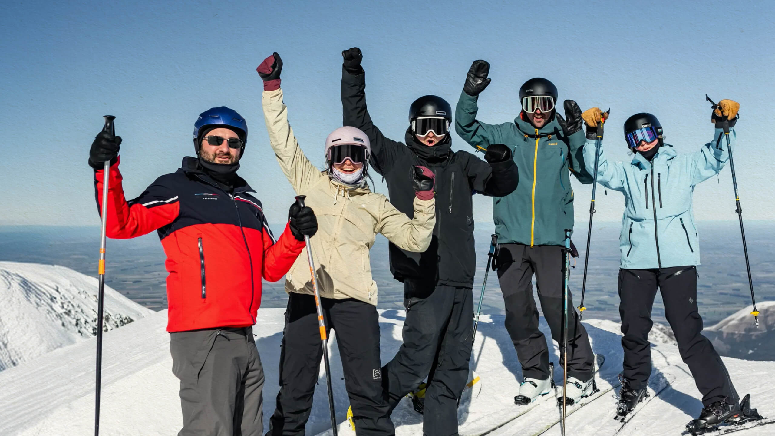 Group of five skiers in helmets and goggles celebrating on a snowy mountain peak at Mt Hutt under a clear blue sky.