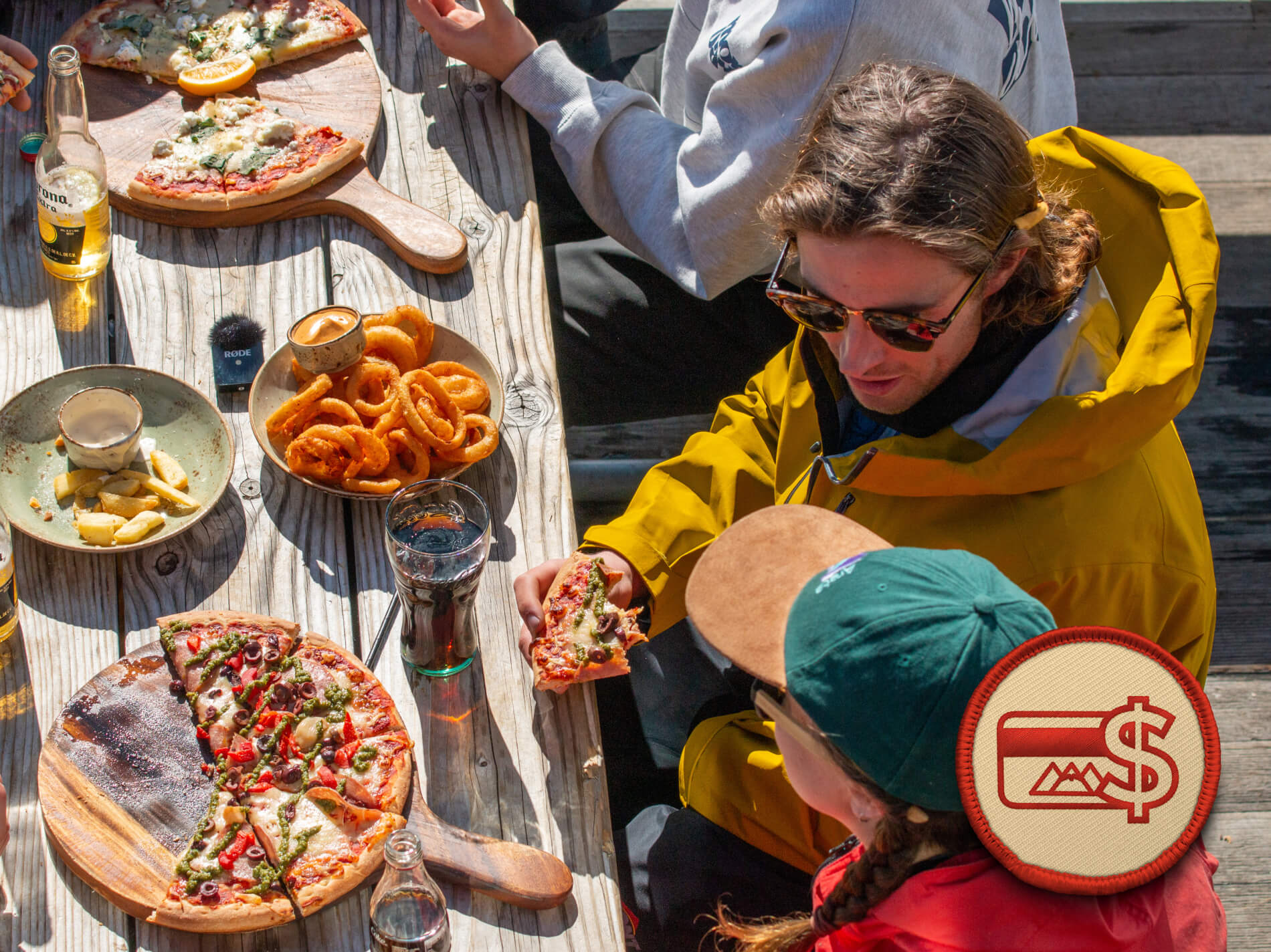 Group of people eating pizza and onion rings at a rustic wooden table with drinks in sunlight.