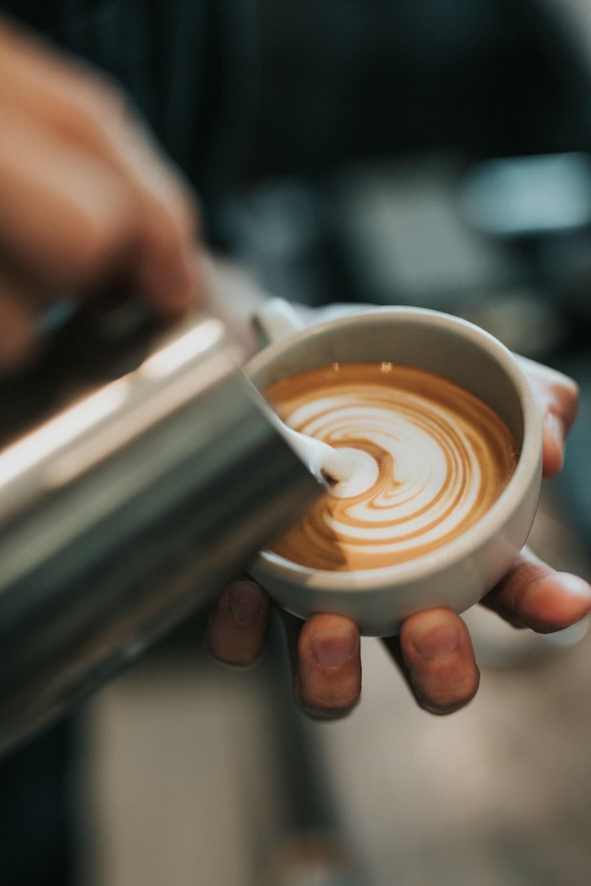 Close-up of a hand pouring milk into a cup of coffee creating latte art swirl.