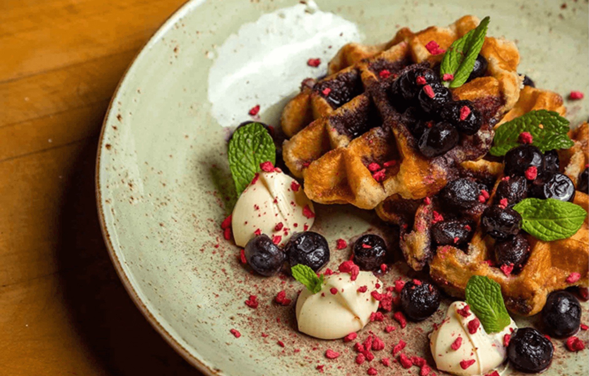 Plate of golden waffles topped with blueberries, mint leaves, cream dollops, and red sprinkles on a light ceramic plate.
