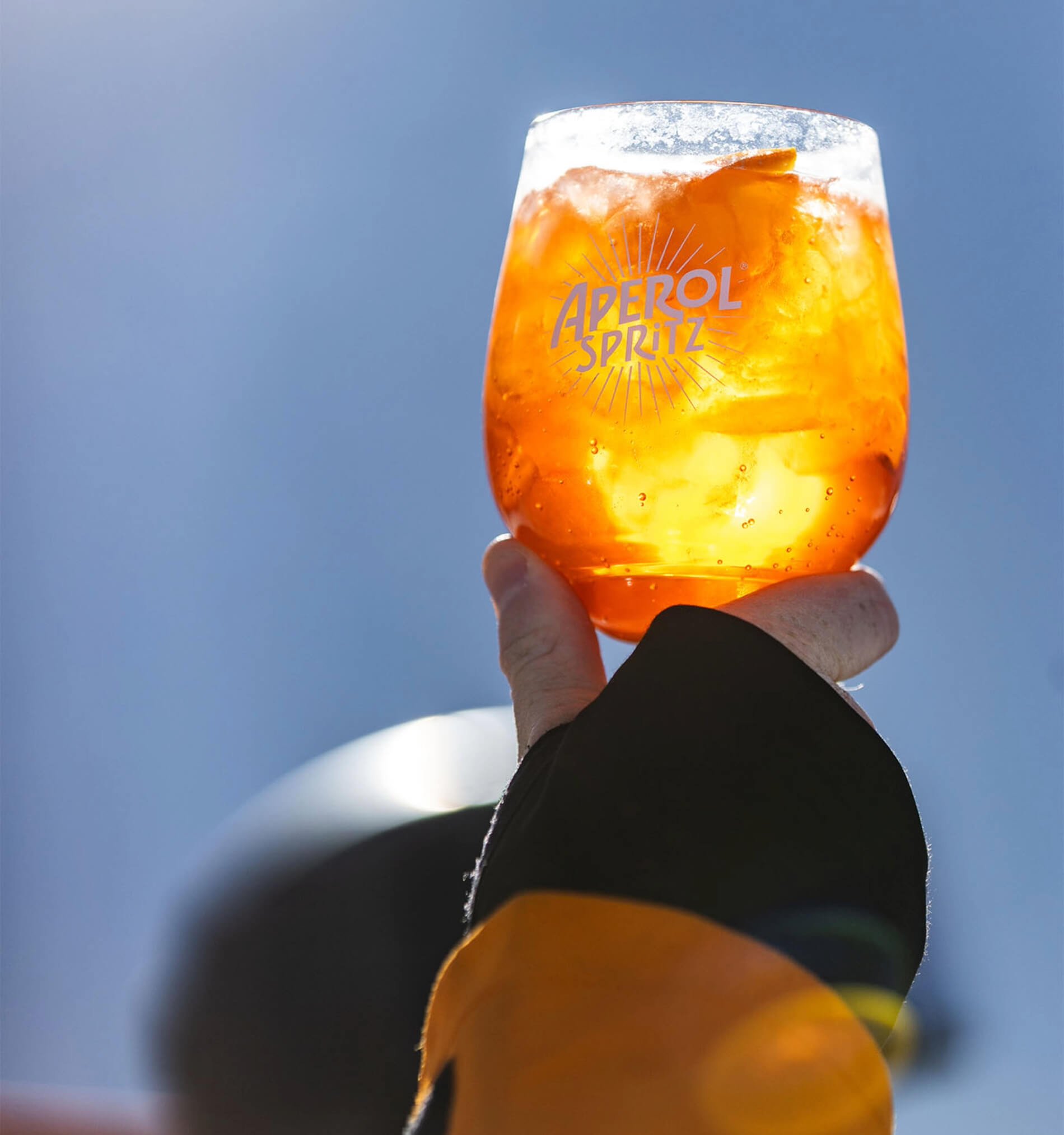 Hand holding a glass of Aperol Spritz with ice and orange slice against a clear blue sky.