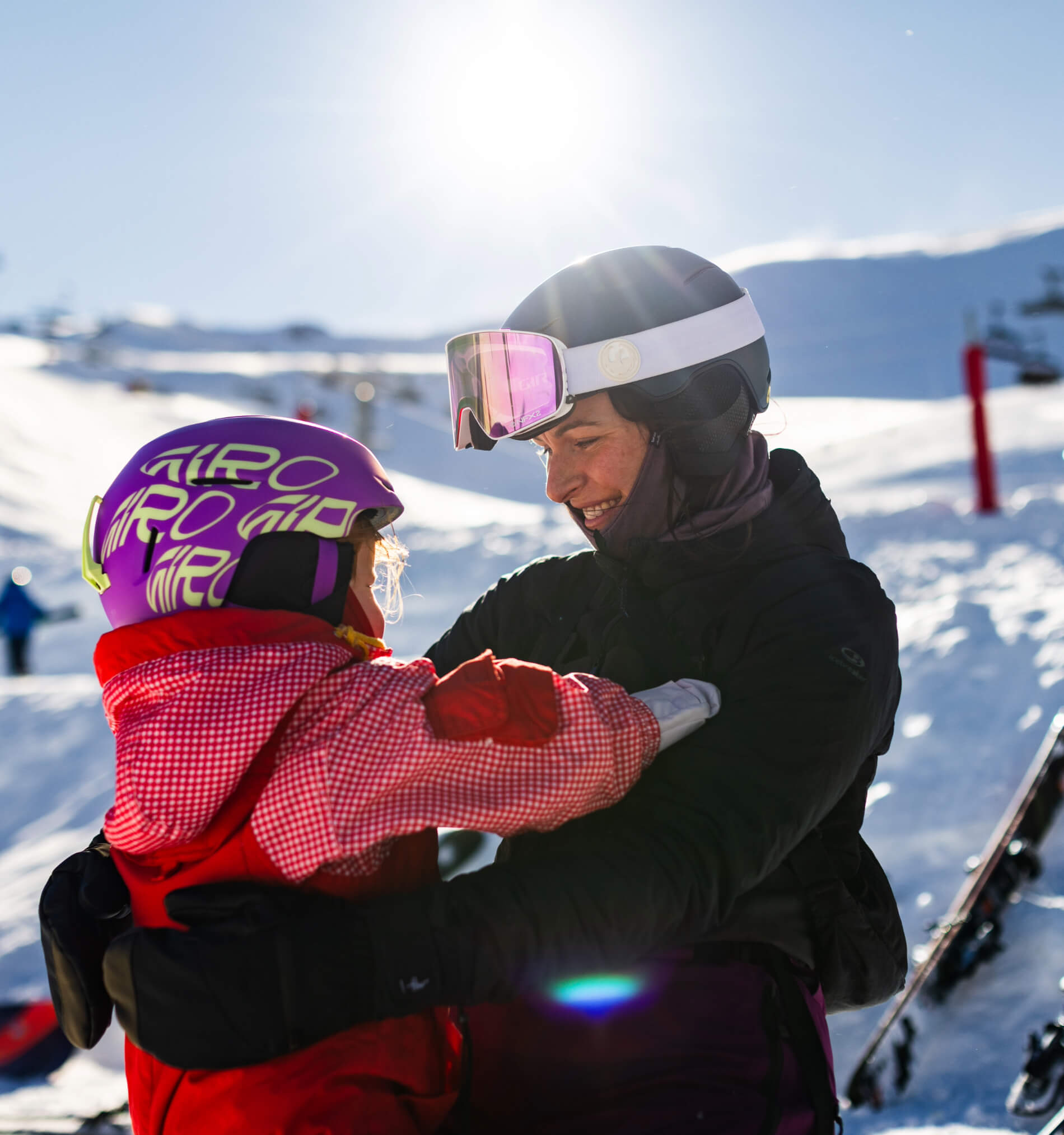 Smiling woman and child wearing ski helmets embracing on a snowy ski slope under bright sun.