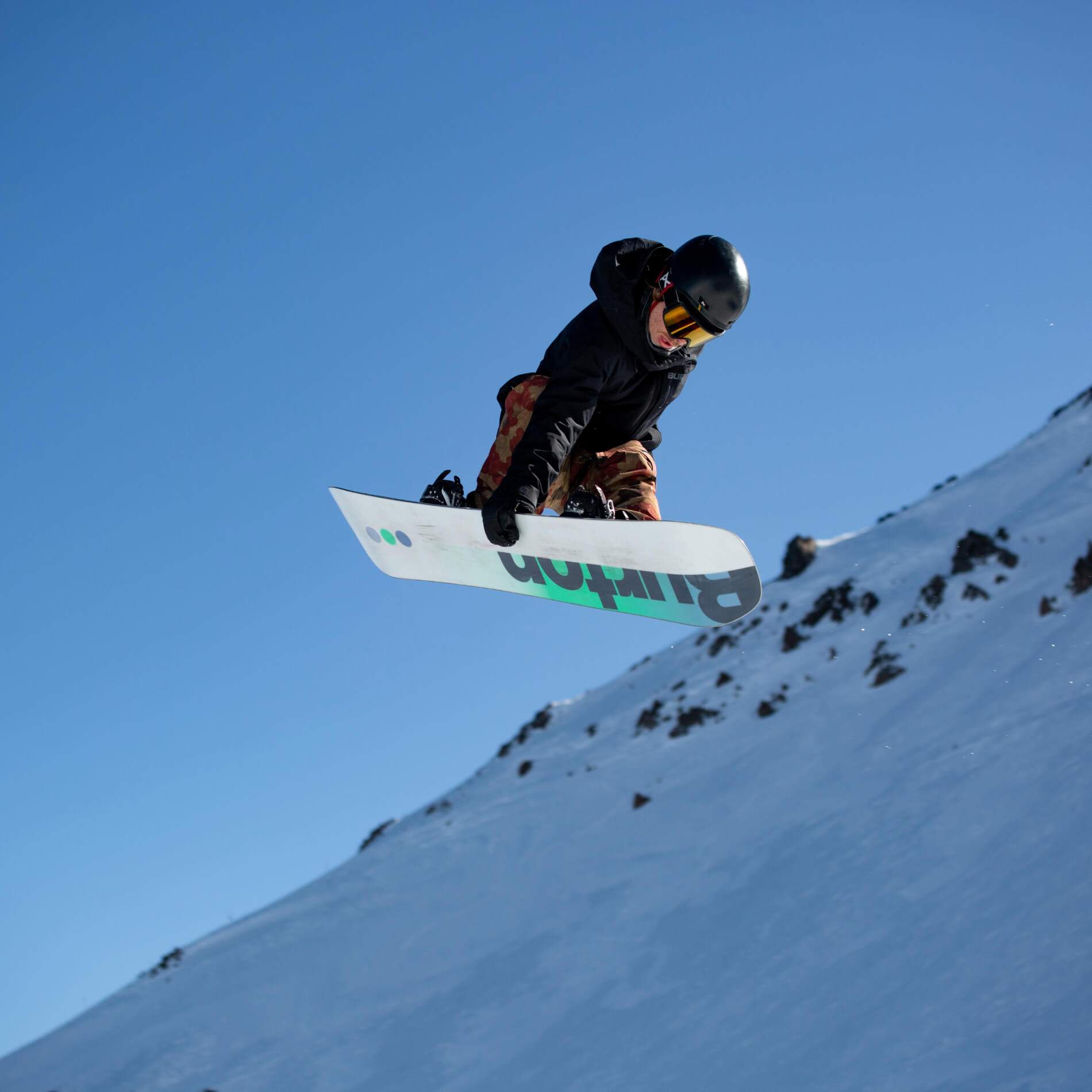 Snowboarder wearing helmet and goggles performs a jump against a clear blue sky and snowy mountain backdrop.