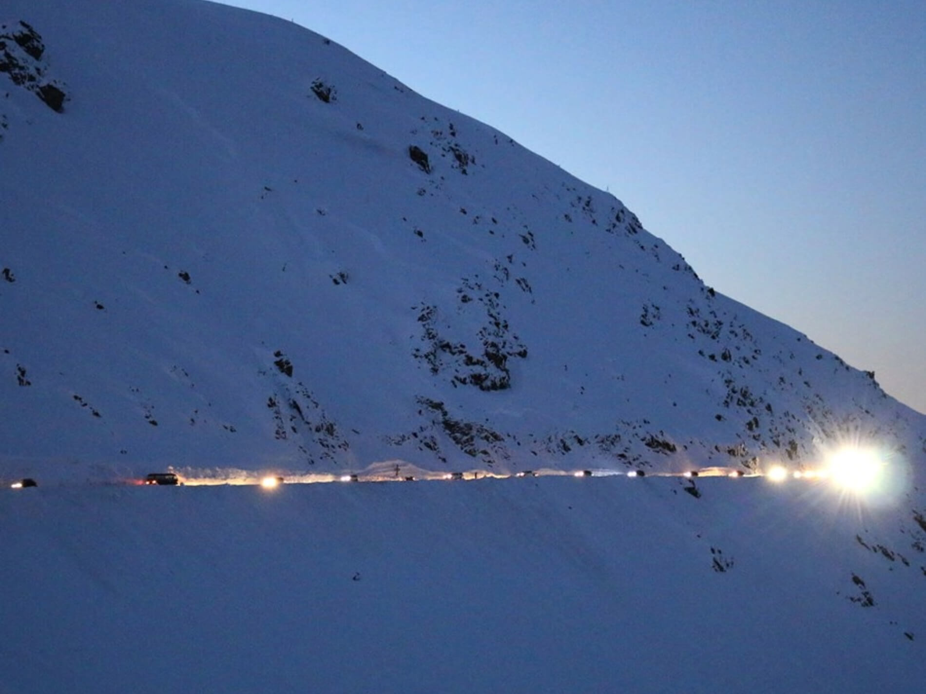 Line of cars with headlights on driving along a snowy mountain road at dusk.
