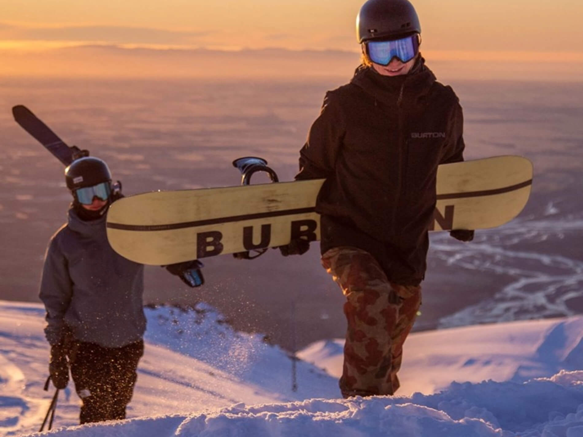 Two snowboarders wearing helmets and goggles walking uphill in snowy mountain terrain at sunset.