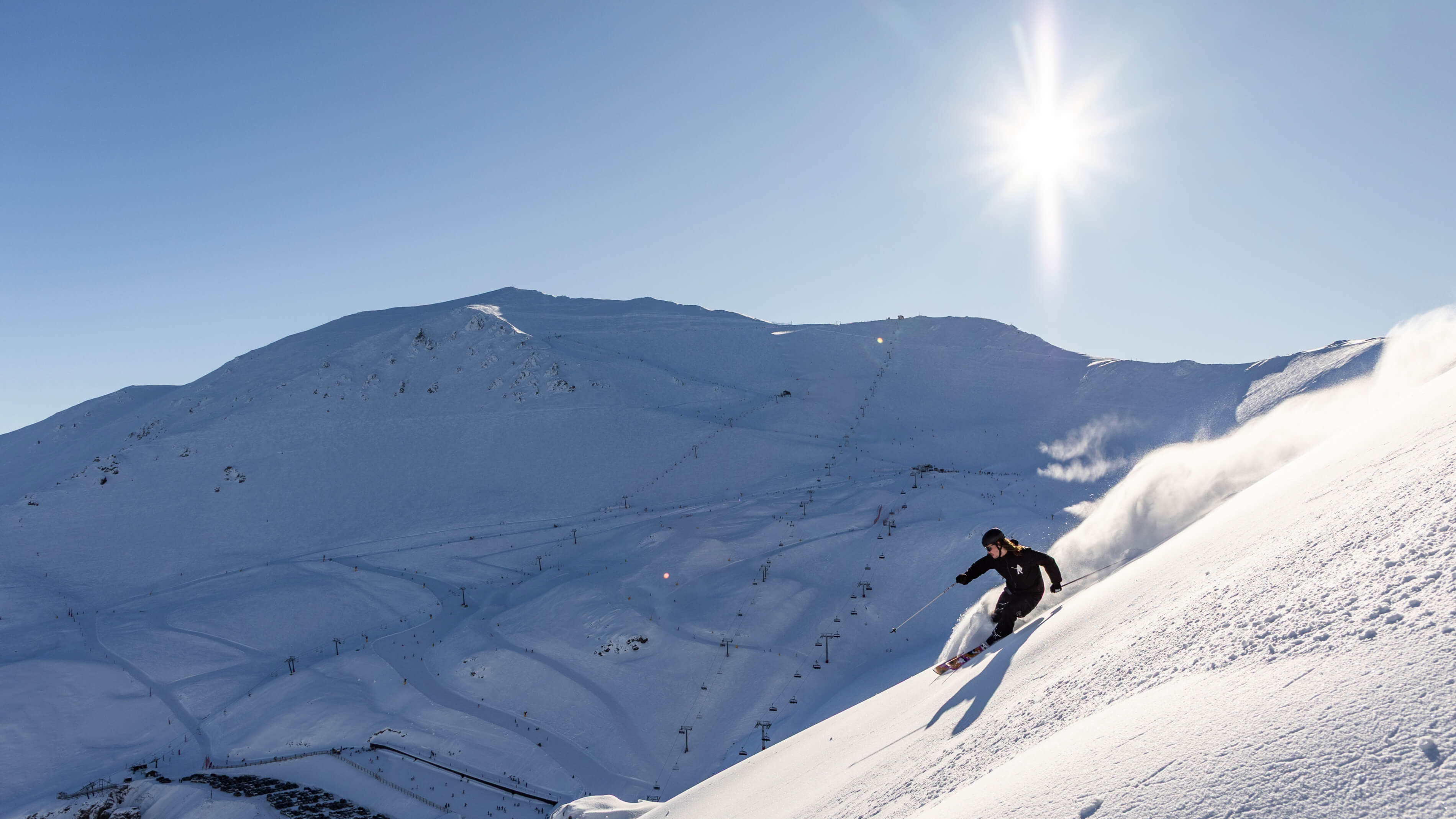 Skier carving down a sunny snowy mountain slope with ski lifts visible on the distant mountain.