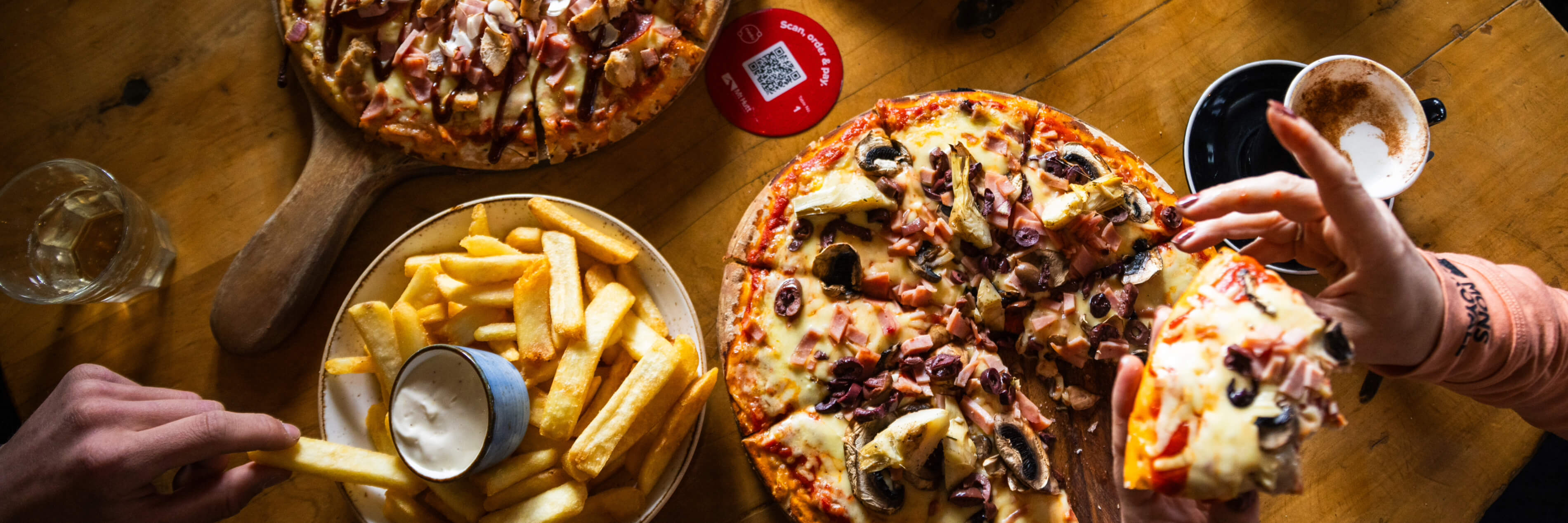 Two hands holding a slice of pizza with a plate of fries and a coffee cup on a wooden table.