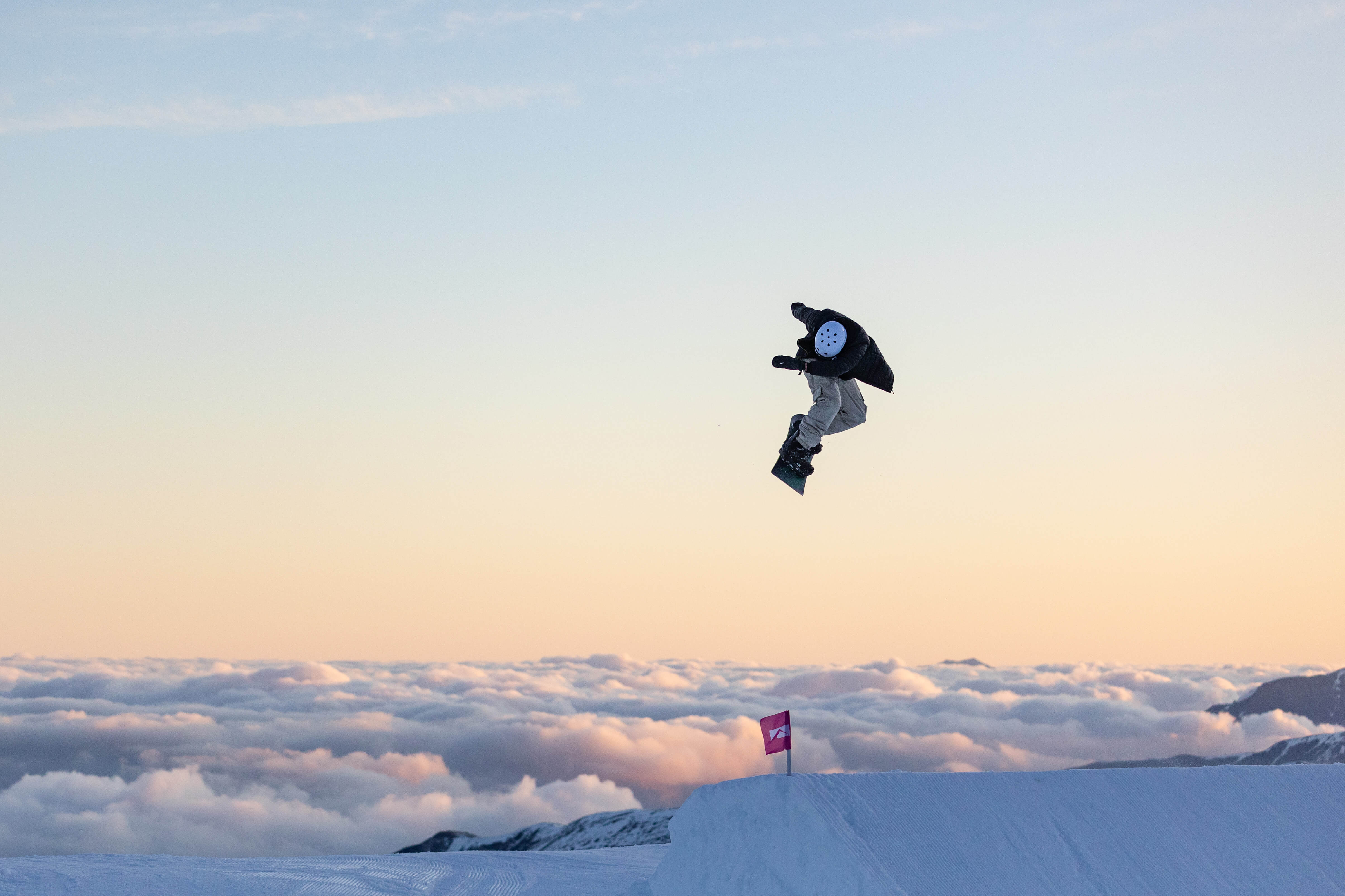 Snowboarder performing a jump over a snow ramp at sunset with clouds and mountains in the background.