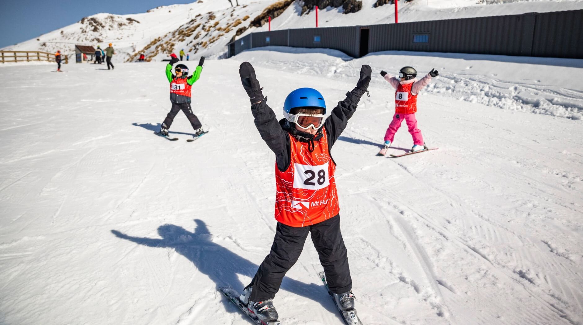 Three children in ski gear with numbered bibs standing on snow with arms raised, practicing skiing on a sunny day.
