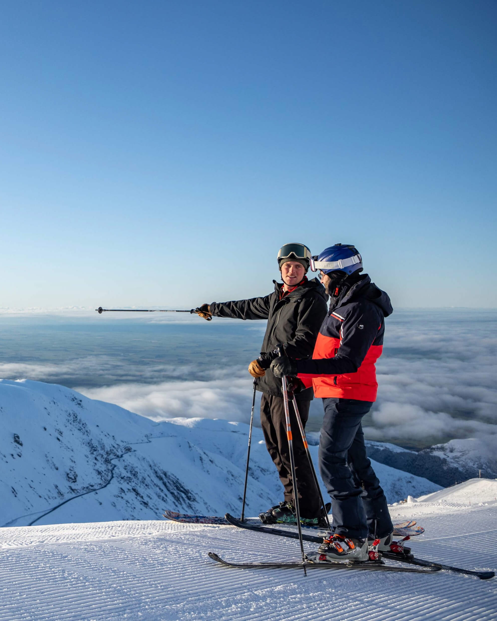 Two skiers standing on a snowy mountain peak above clouds, one pointing into the distance under a clear blue sky.