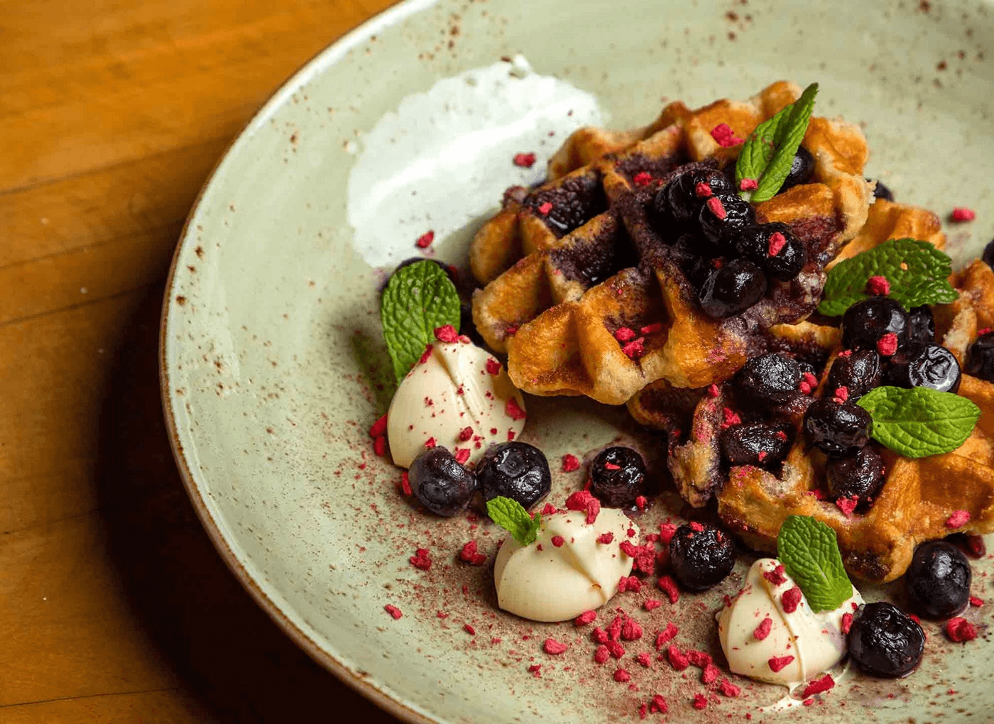 Plate of waffles topped with blueberries, mint leaves, dollops of cream, and sprinkled with red crumbs.