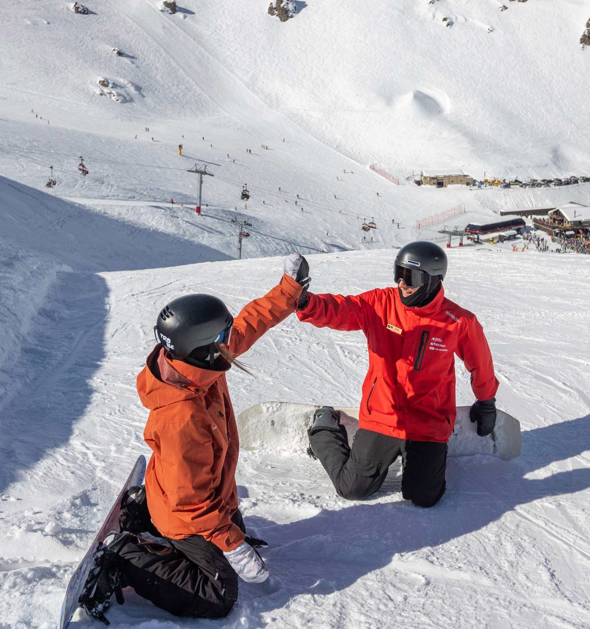 Two snowboarders kneeling on snow giving each other a high five with snowy mountain and ski lifts in the background.