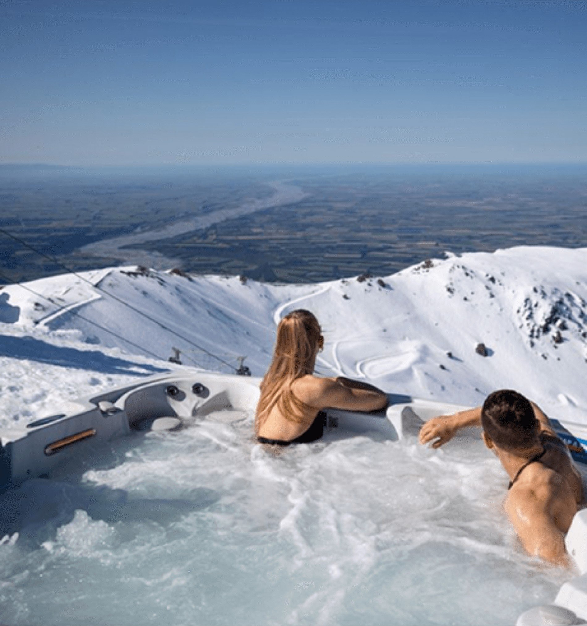 Couple relaxing in an outdoor hot tub overlooking snow-covered mountains and a valley below under a clear blue sky.