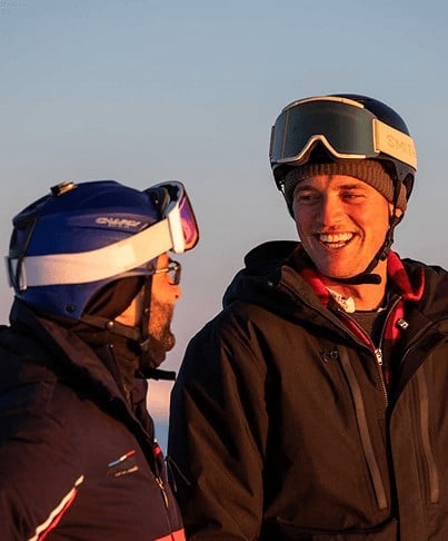 Two men wearing ski helmets and jackets smiling and talking outdoors at sunset.
