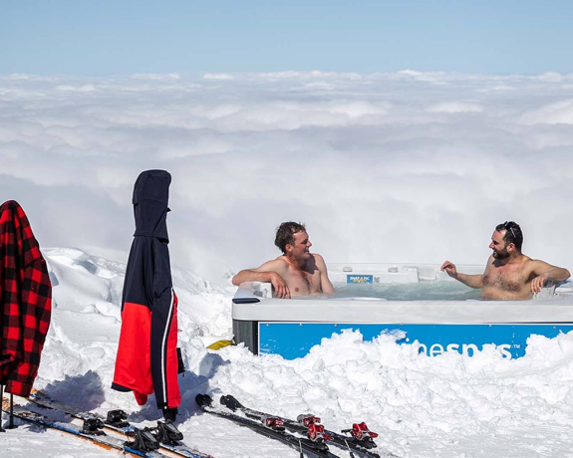 Two men relaxing and chatting in the Mt Hutt Alpine Spa surrounded by snow with ski gear and jackets hanging nearby.