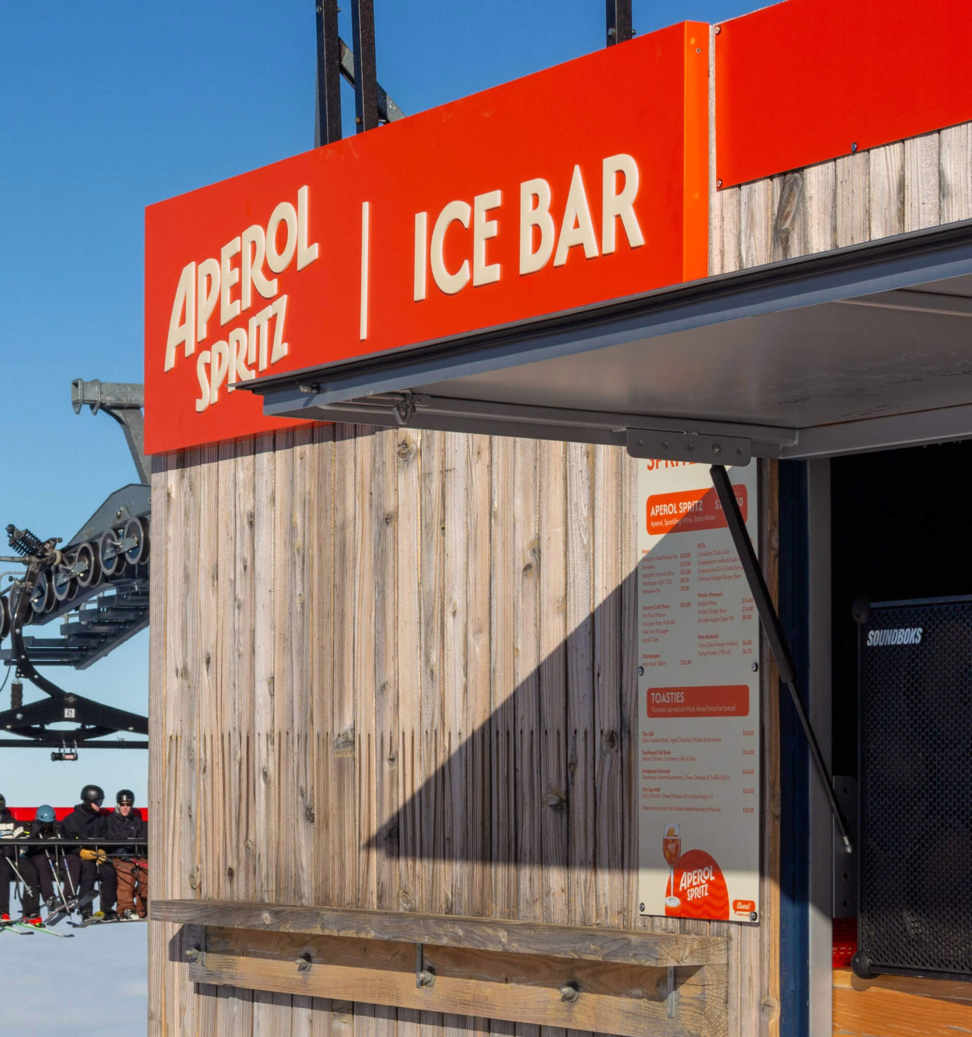 Wood-paneled Aperol Spritz Ice Bar kiosk with menu and ski lift in the background under clear blue sky.