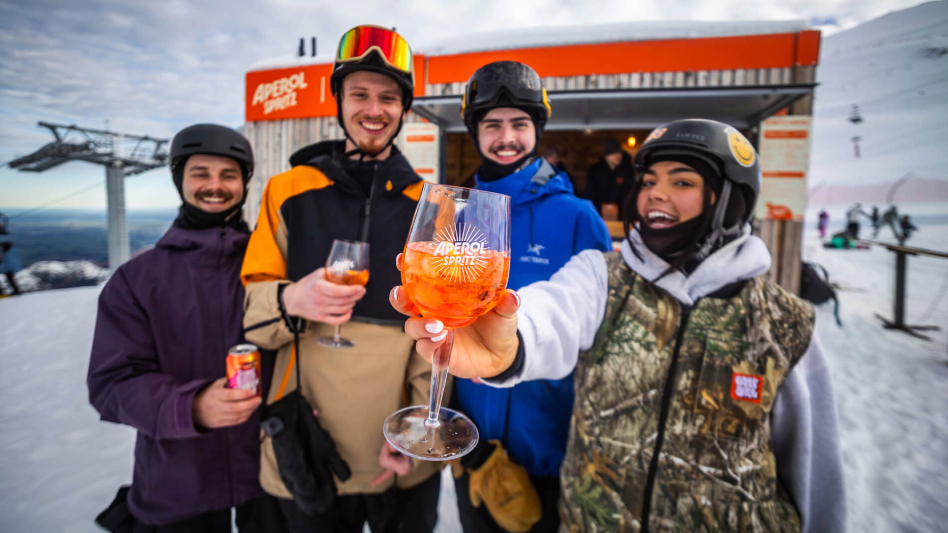 Four friends in ski gear holding Aperol Spritz drinks in a snowy mountain setting with an Aperol Spritz bar in the background.