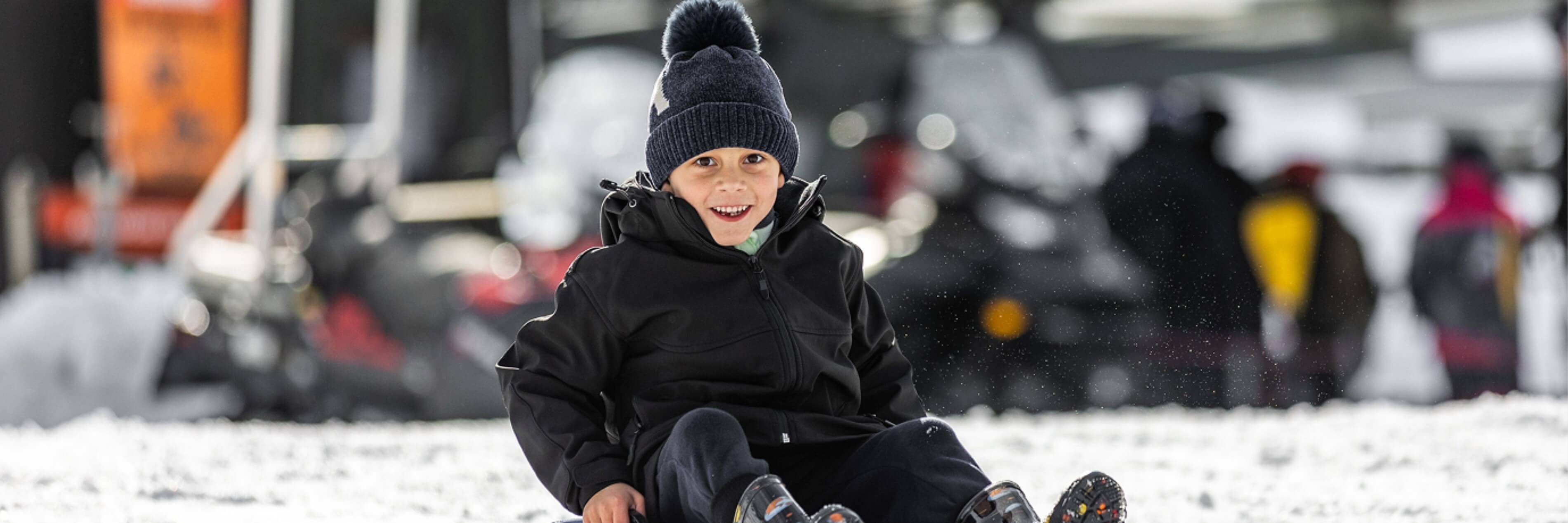Smiling boy in a black jacket and beanie sledding on snow with blurred people and vehicles in the background.