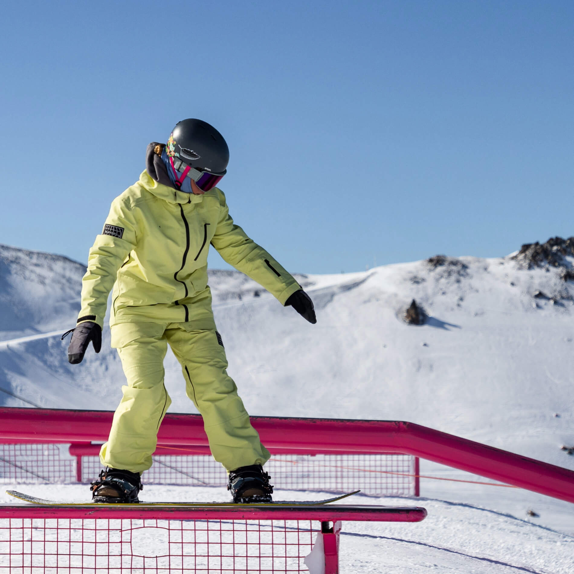 Snowboarder in yellow jacket and pants sliding on a pink rail with snowy mountains in the background.