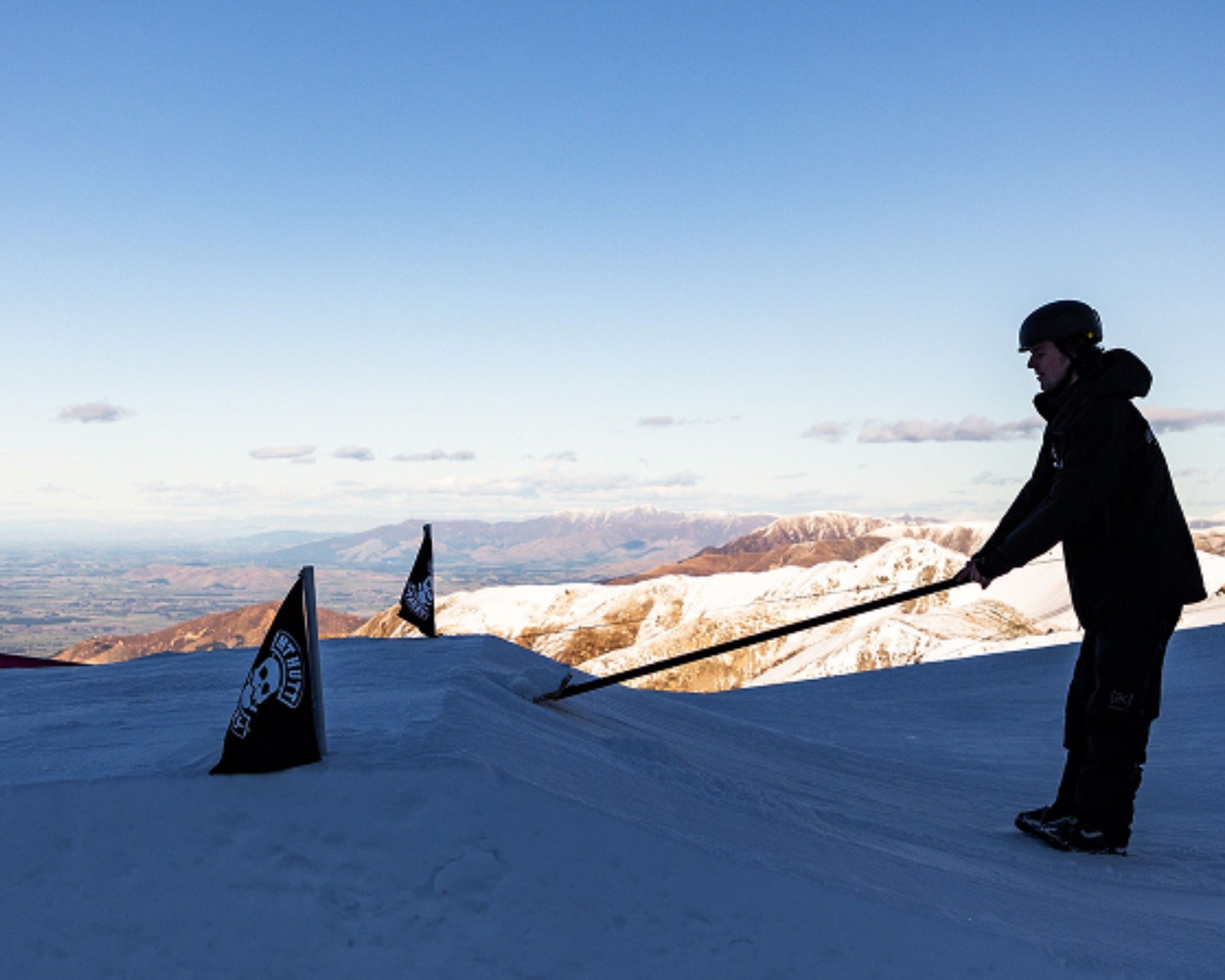 Person wearing a helmet grooming snow on a ski slope with a tool near black flags on a mountain under a clear sky.