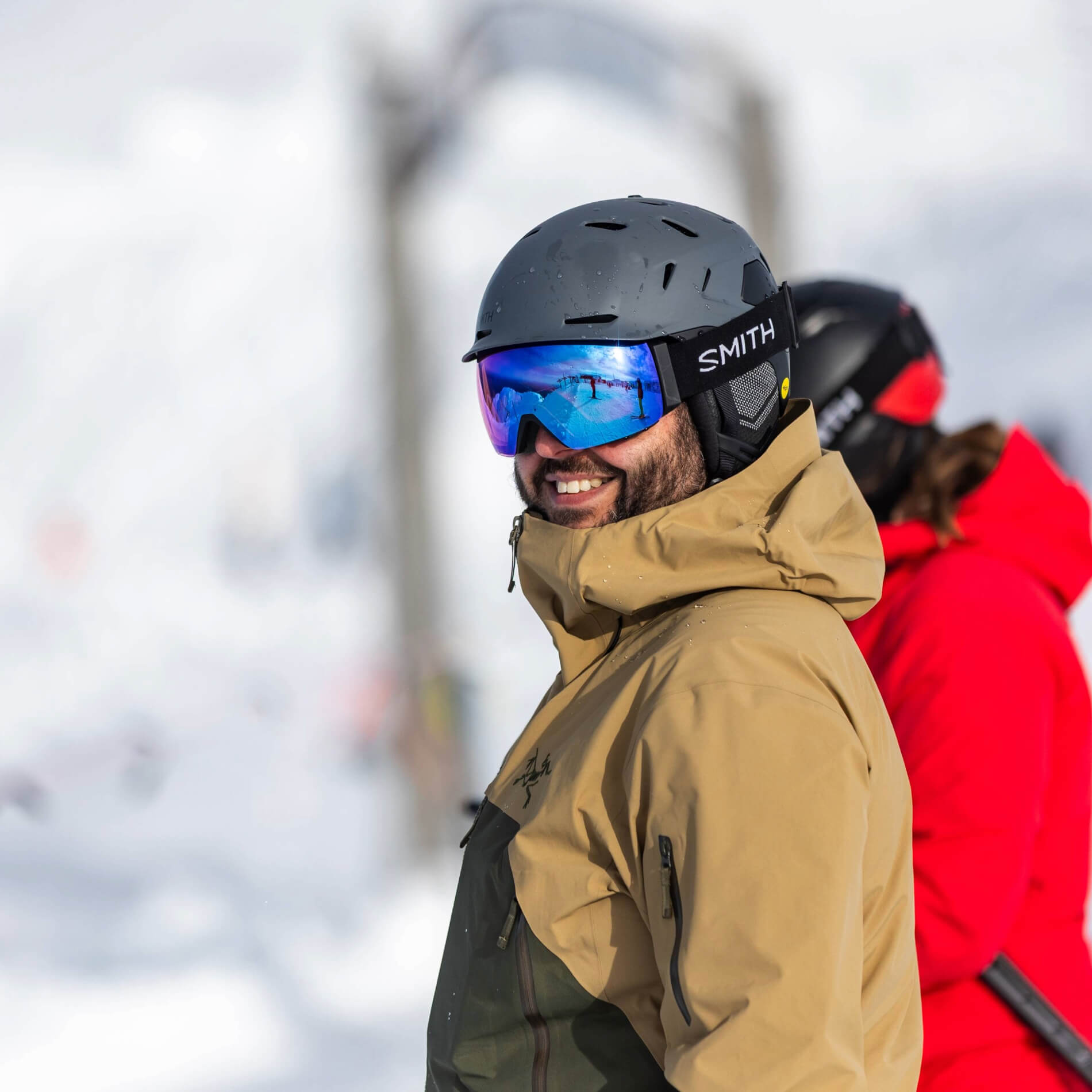 Smiling man wearing a black helmet and reflective ski goggles with a tan and green winter jacket, standing in snowy mountain gear with another person in red in the background.