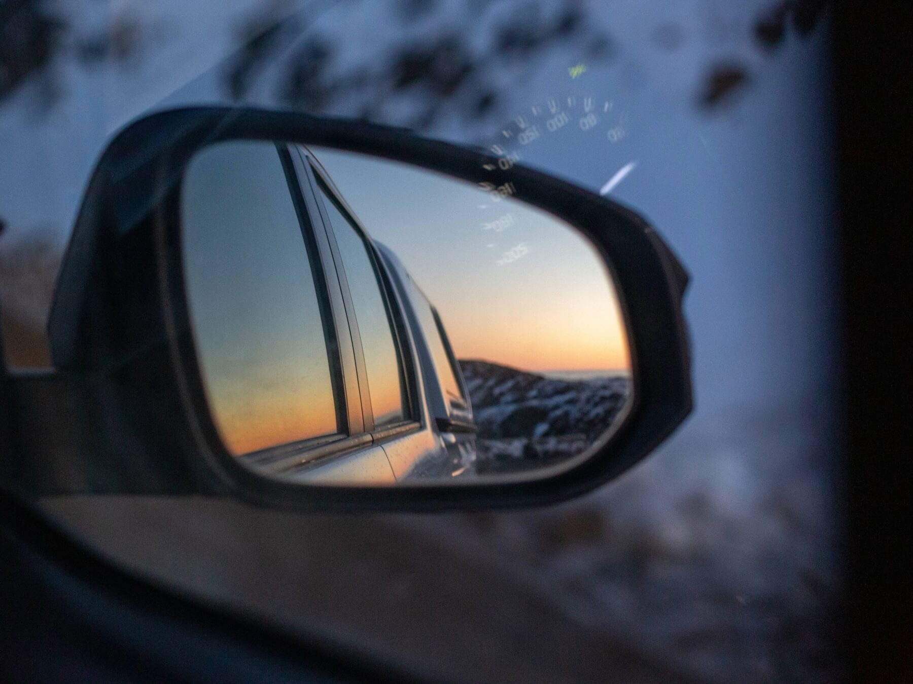 Reflection of a sunset sky and rocky landscape in a car's side mirror.