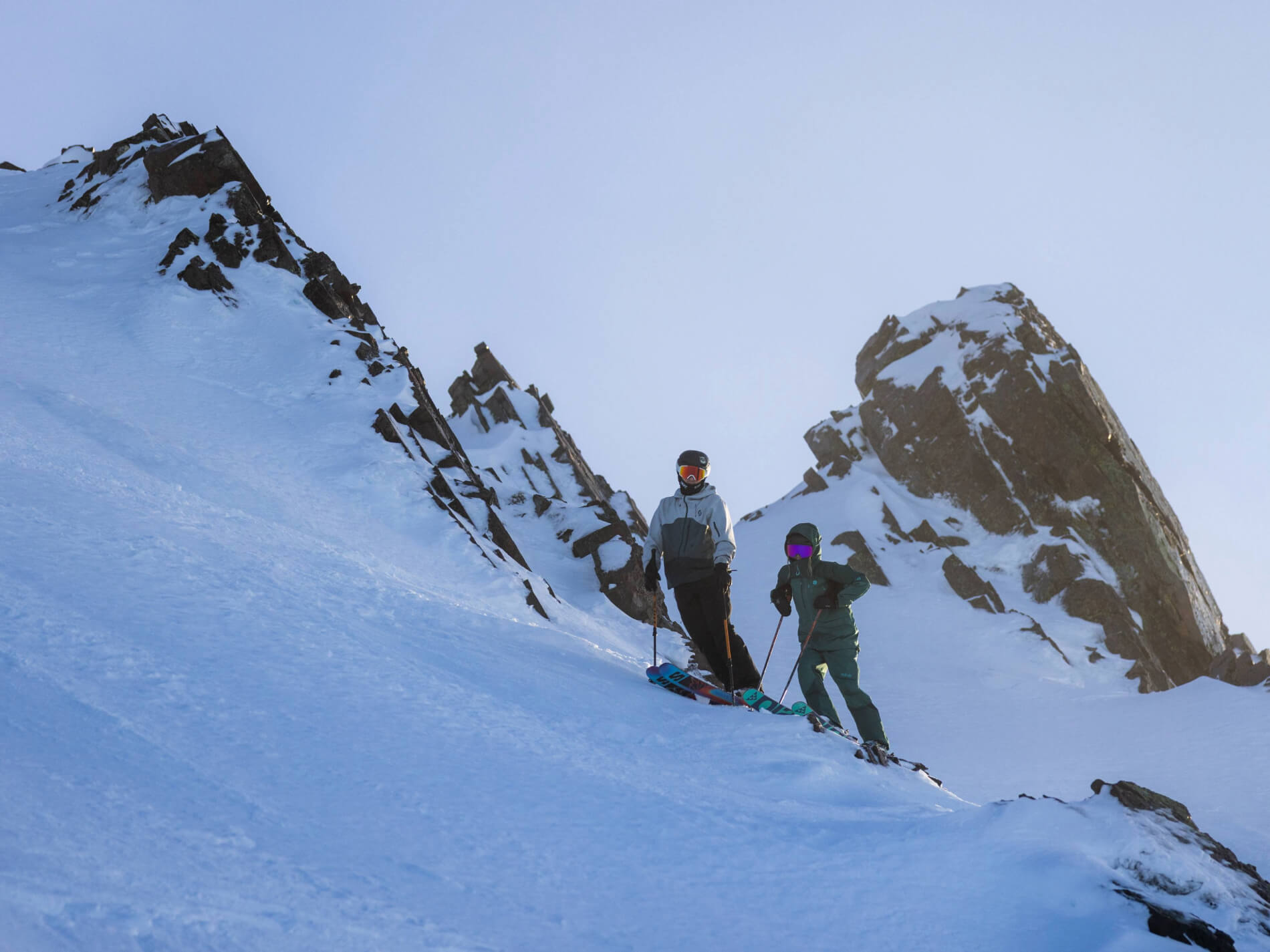 Two skiers standing on a snowy mountain slope with rocky peaks in the background under a clear sky.