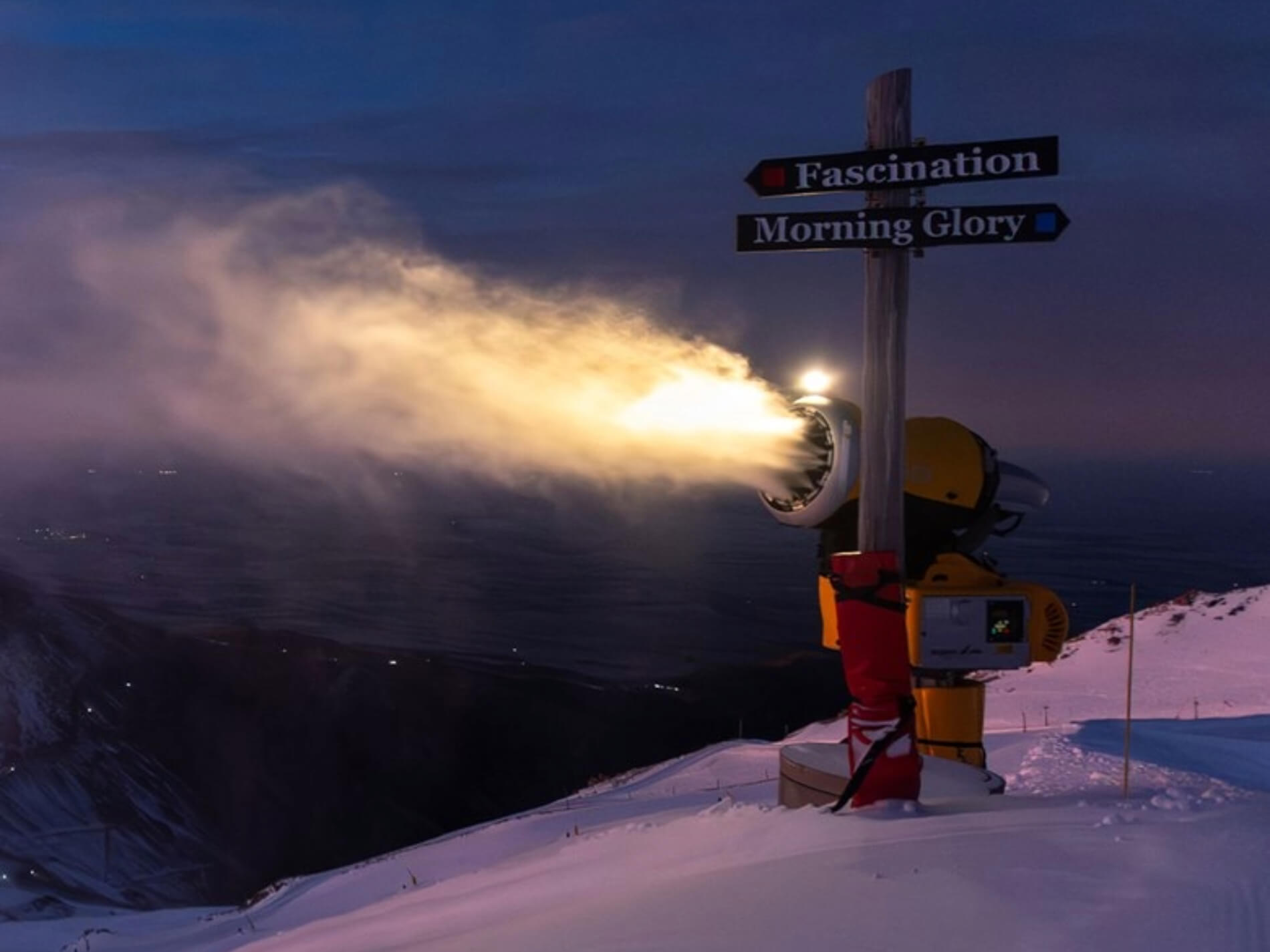 Snow machine blowing snow at night on Mt Hutt near a signpost with directions labeled Fascination and Morning Glory.