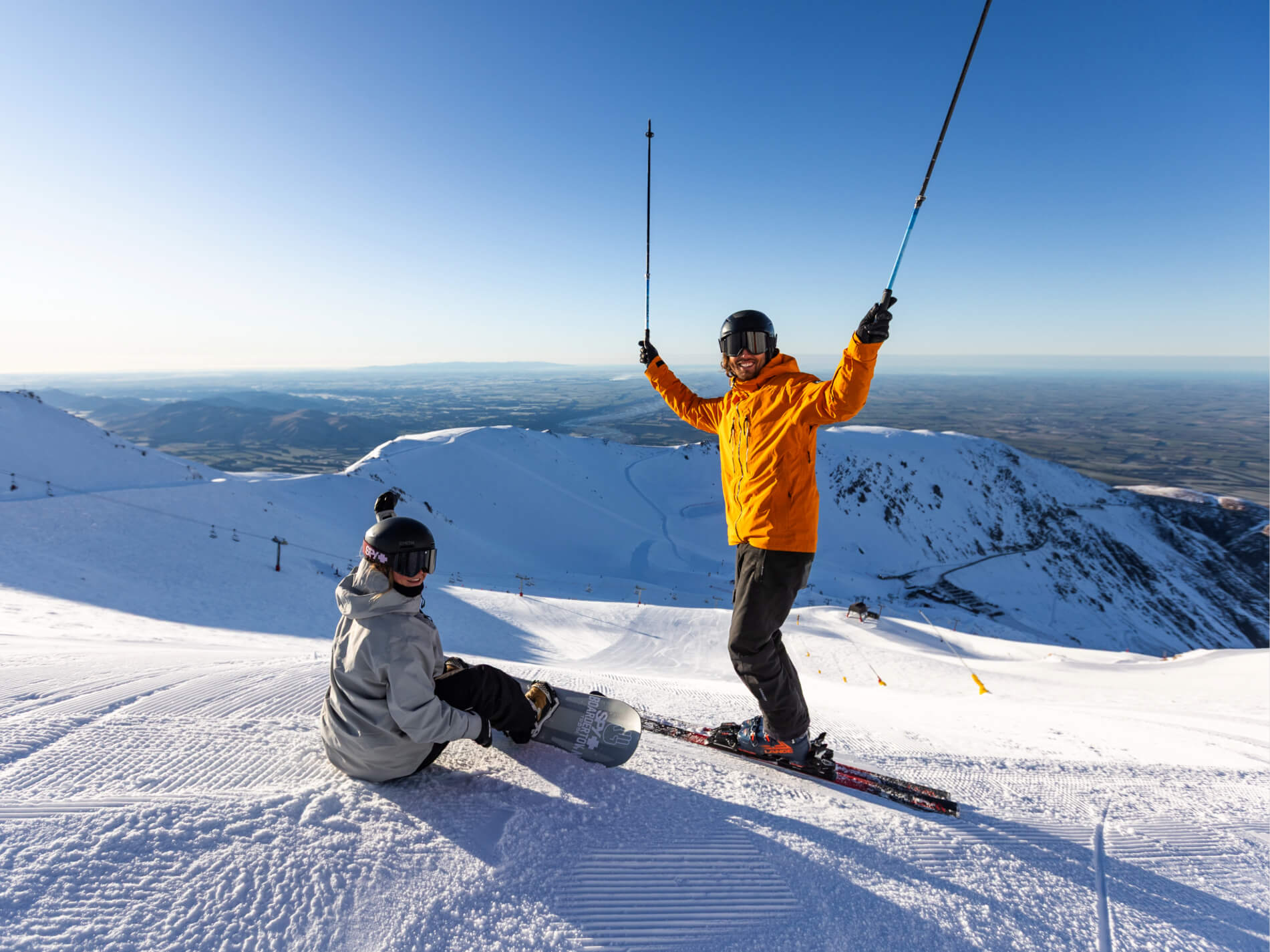 Smiling skier in an orange jacket holding ski poles standing next to a seated snowboarder in a gray jacket on a snowy mountain slope under a clear blue sky.