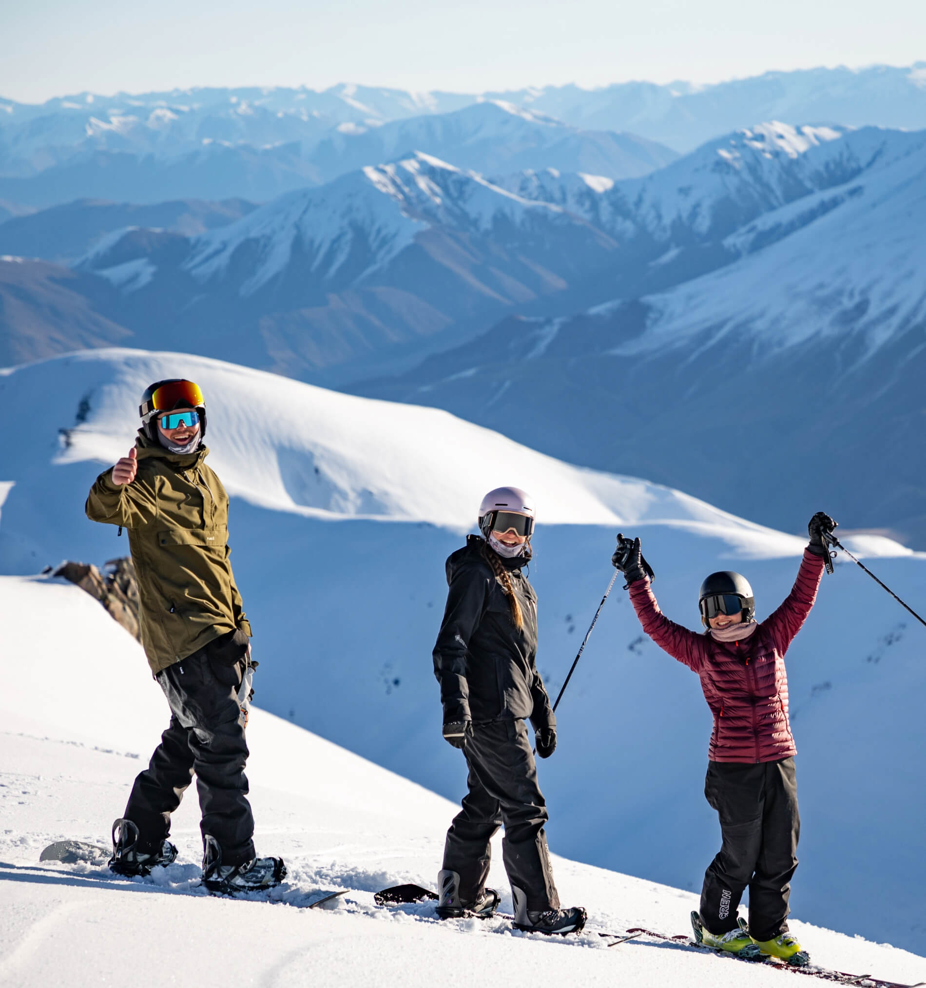 Three snowboarders and skiers on snowy mountain with snow-covered peaks in the background, one giving a thumbs up.