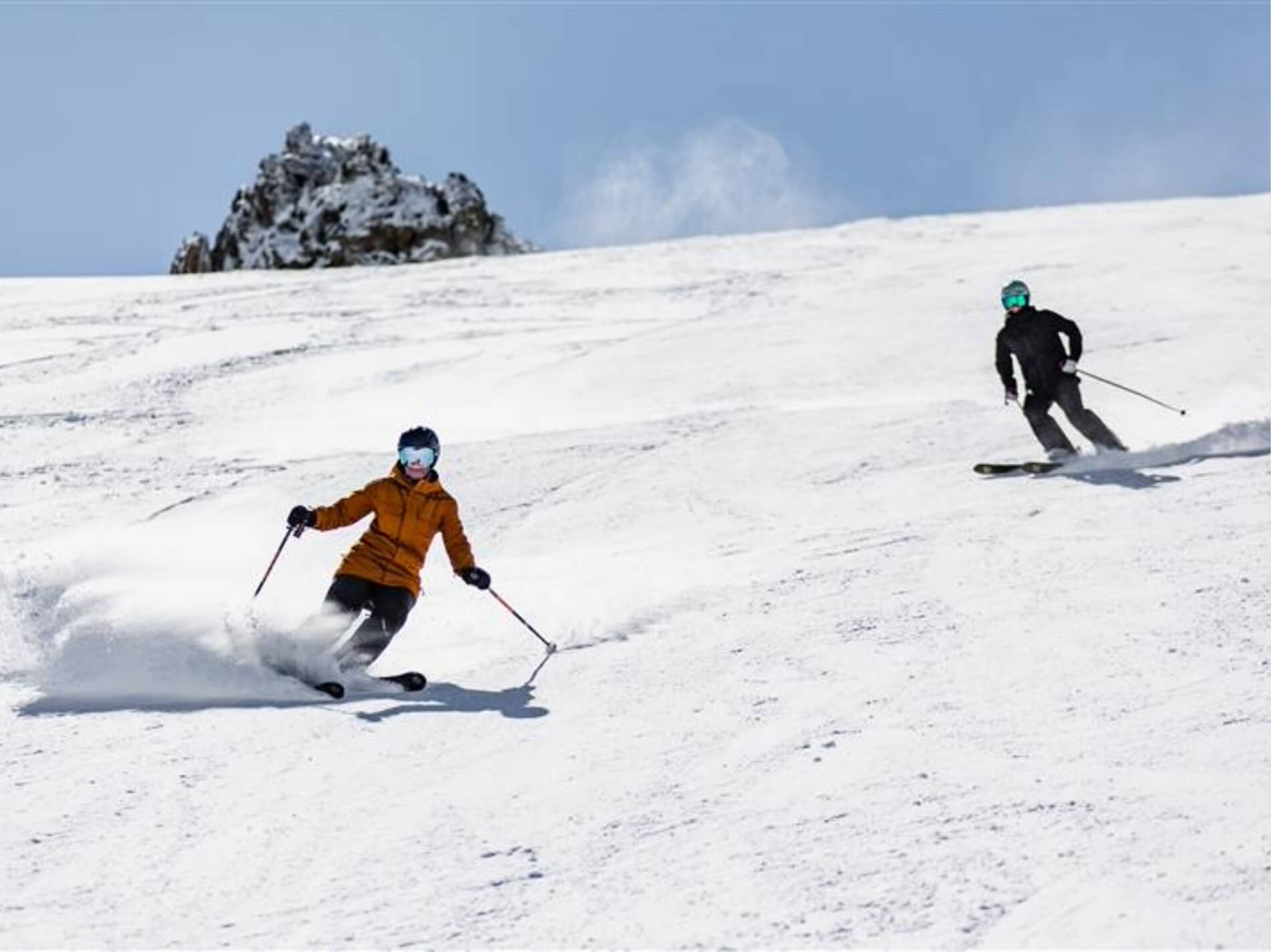Two skiers in winter gear skiing downhill on a snowy slope under a clear blue sky.
