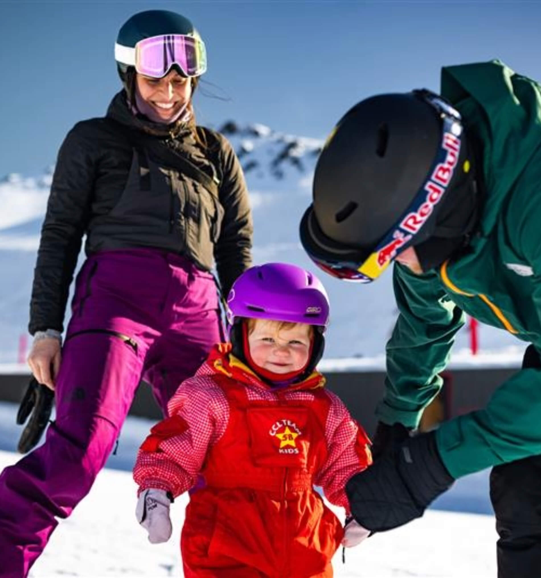 Toddler wearing a purple helmet and red ski outfit smiling between two adults on a snowy mountain.