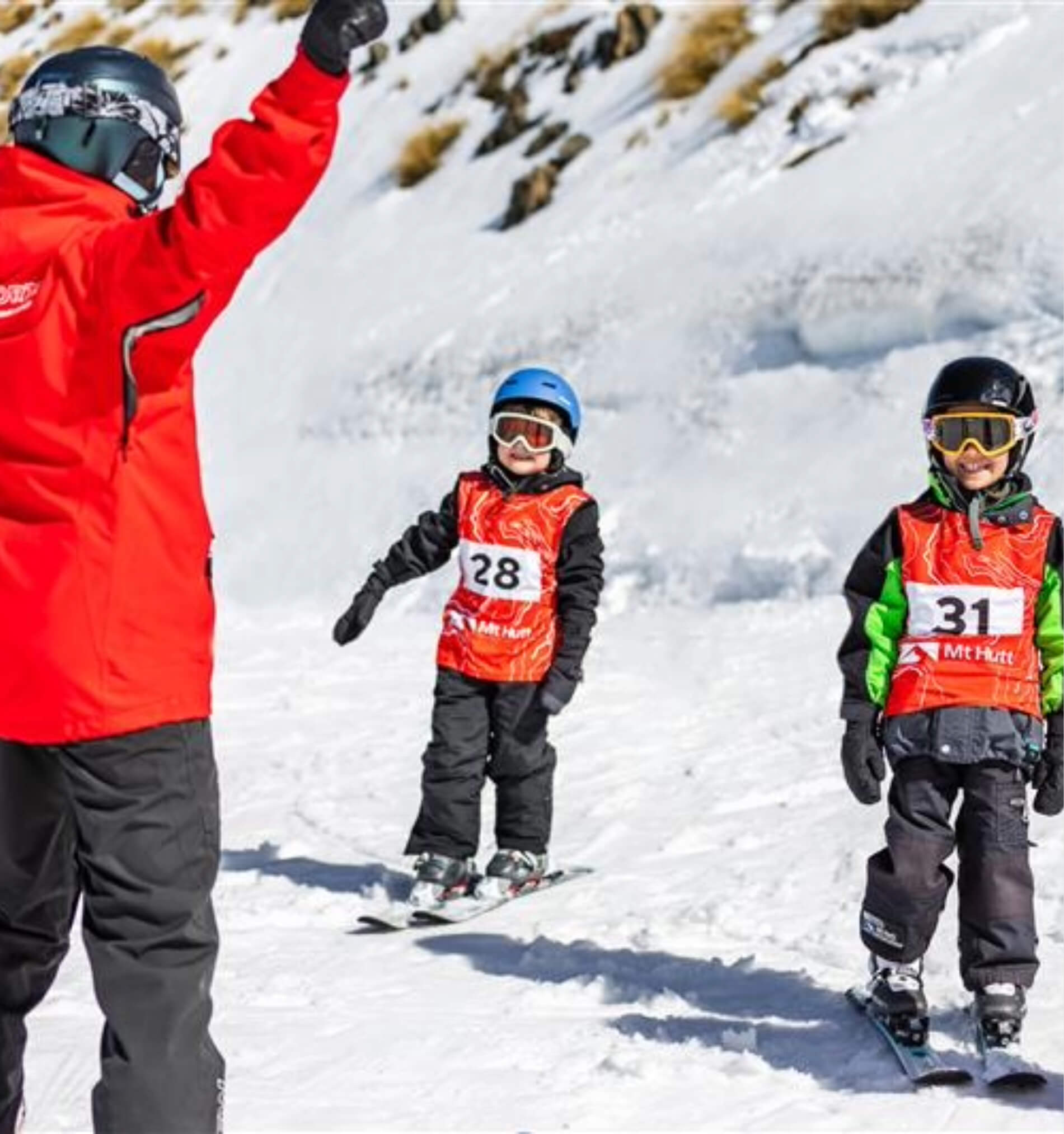 Two children wearing helmets and numbered ski vests learning to ski on snow with an instructor in a red jacket.