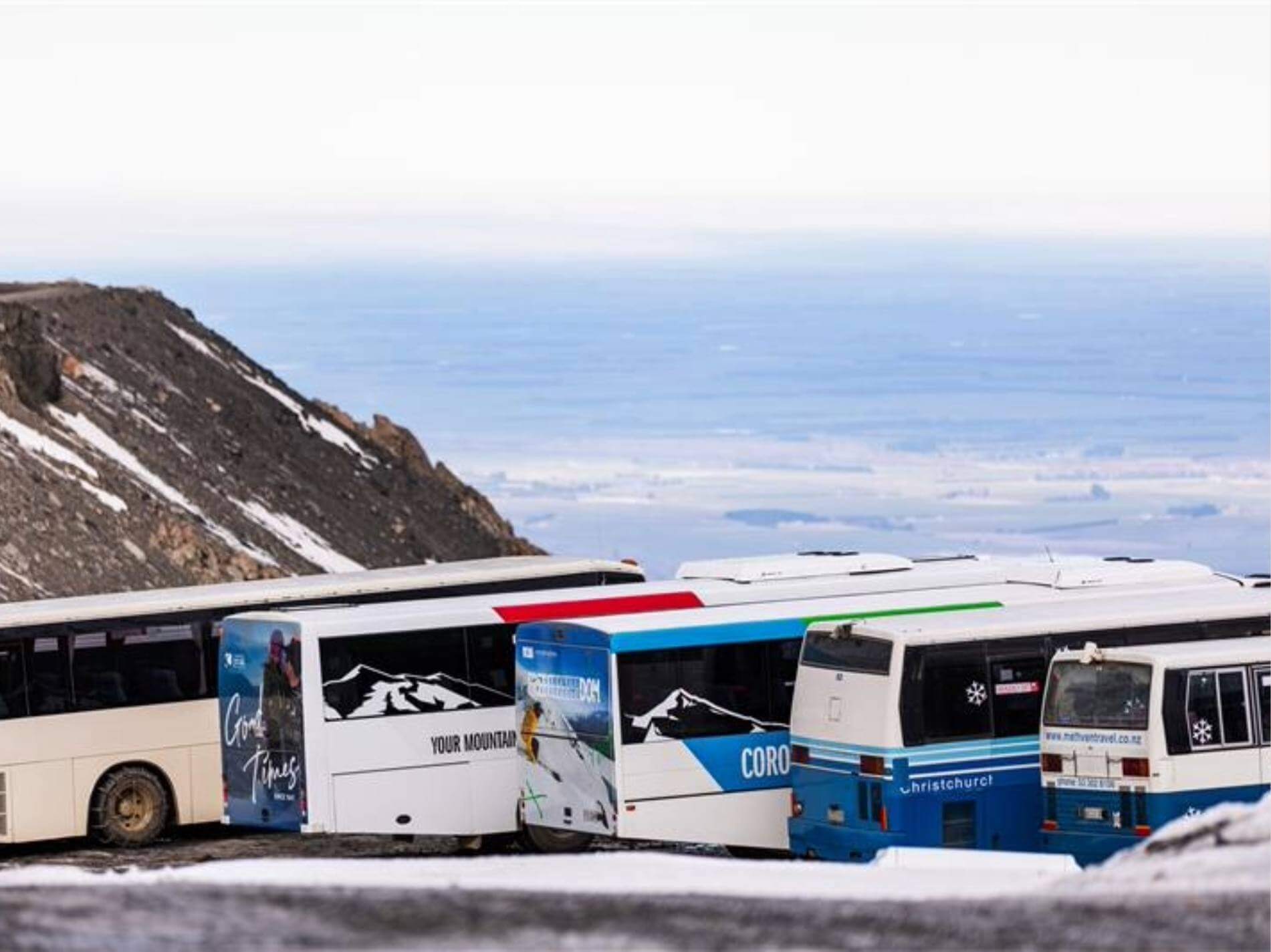 Row of parked buses with mountain and snow background, overlooking a distant valley view.