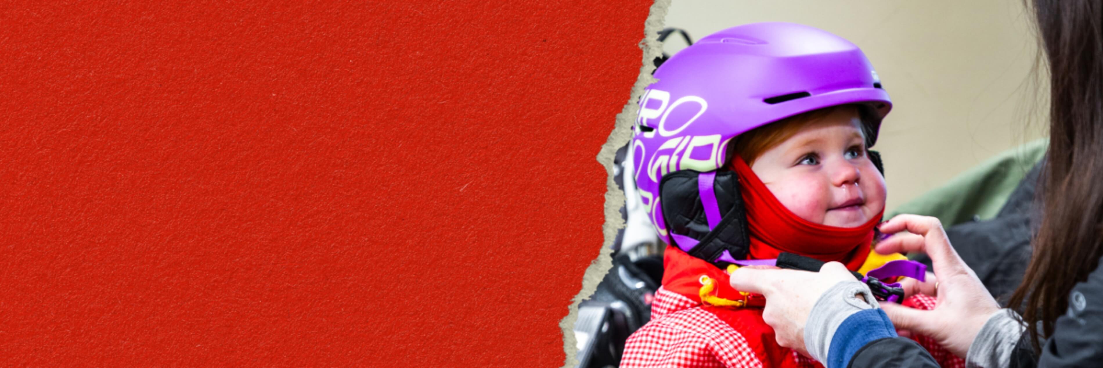 Toddler in a purple helmet and red jacket looking up while an adult secures her helmet strap.