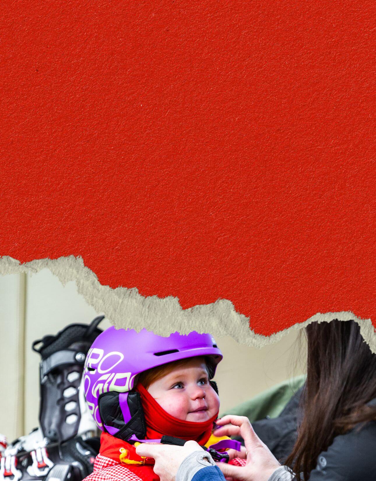 Toddler wearing a purple helmet and red winter clothes, being helped with her helmet strap by an adult.