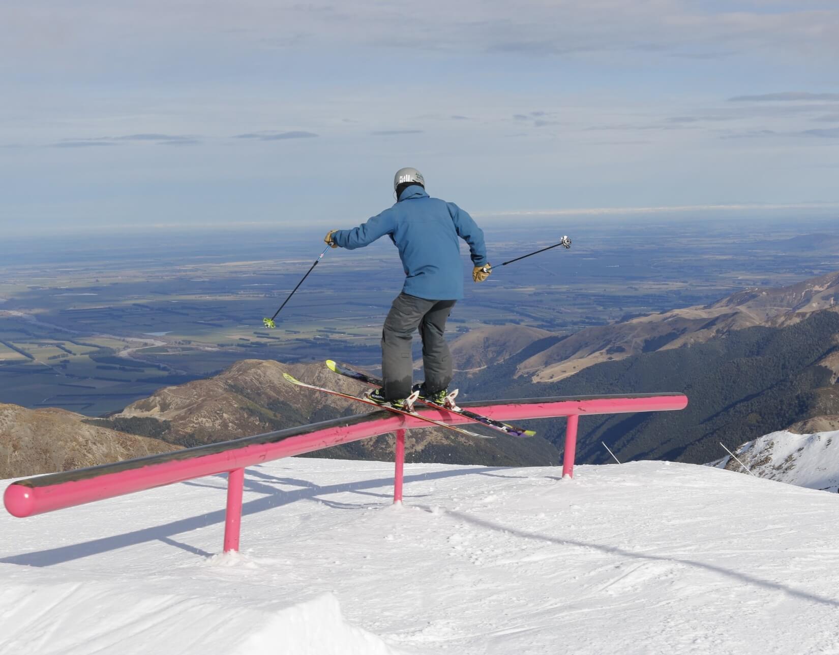Skier in blue jacket sliding on a pink rail on a snowy mountain with a valley and mountains in the background.