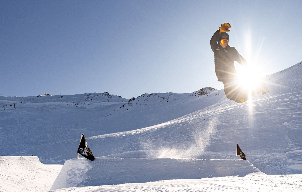 Snowboarder wearing a black outfit and orange gloves performing a jump on a snow terrain park with sun glare behind.