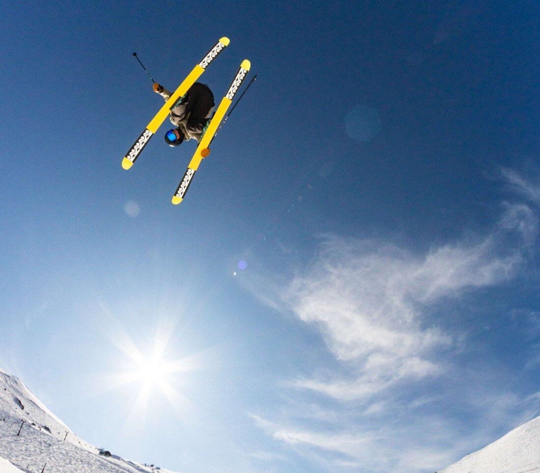 Skier wearing a blue helmet performing a jump in the air against a clear blue sky with the sun shining and snowy slopes below.