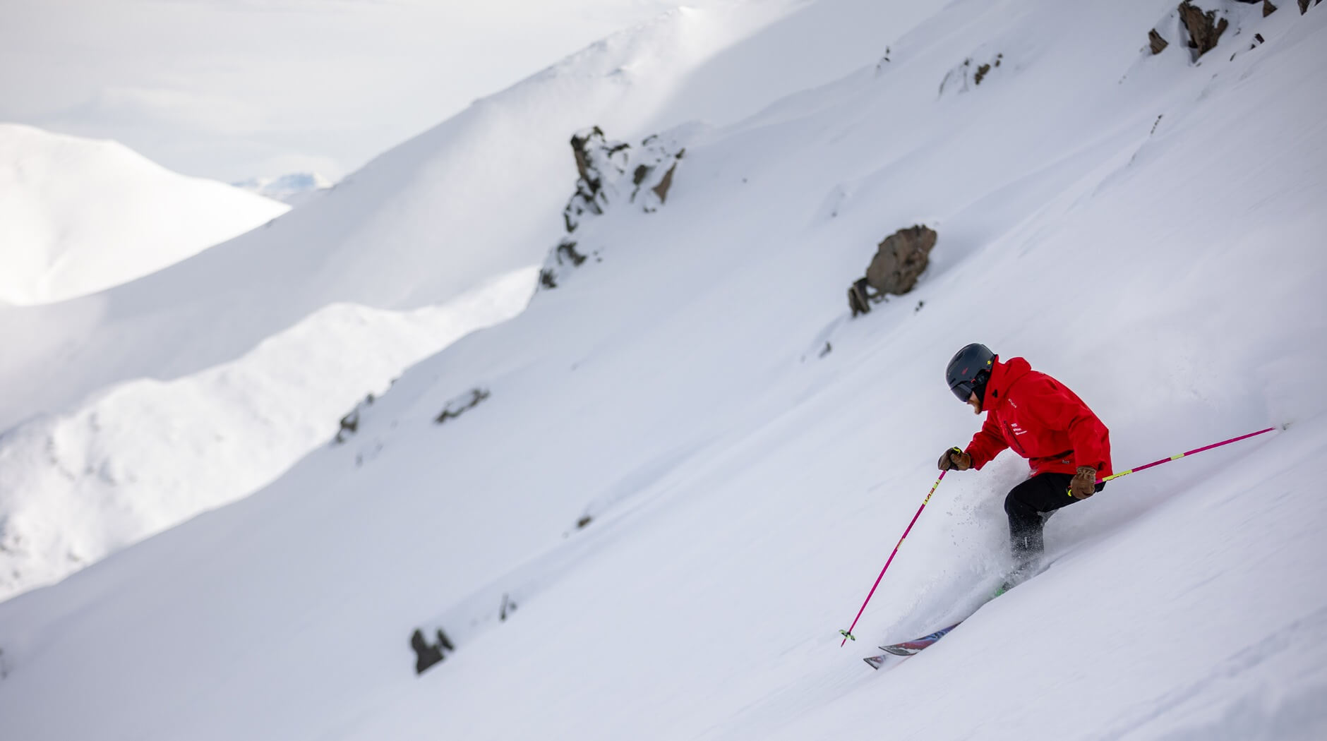 Skier in a red jacket and black helmet skiing down a snowy mountain slope with scattered rocks.