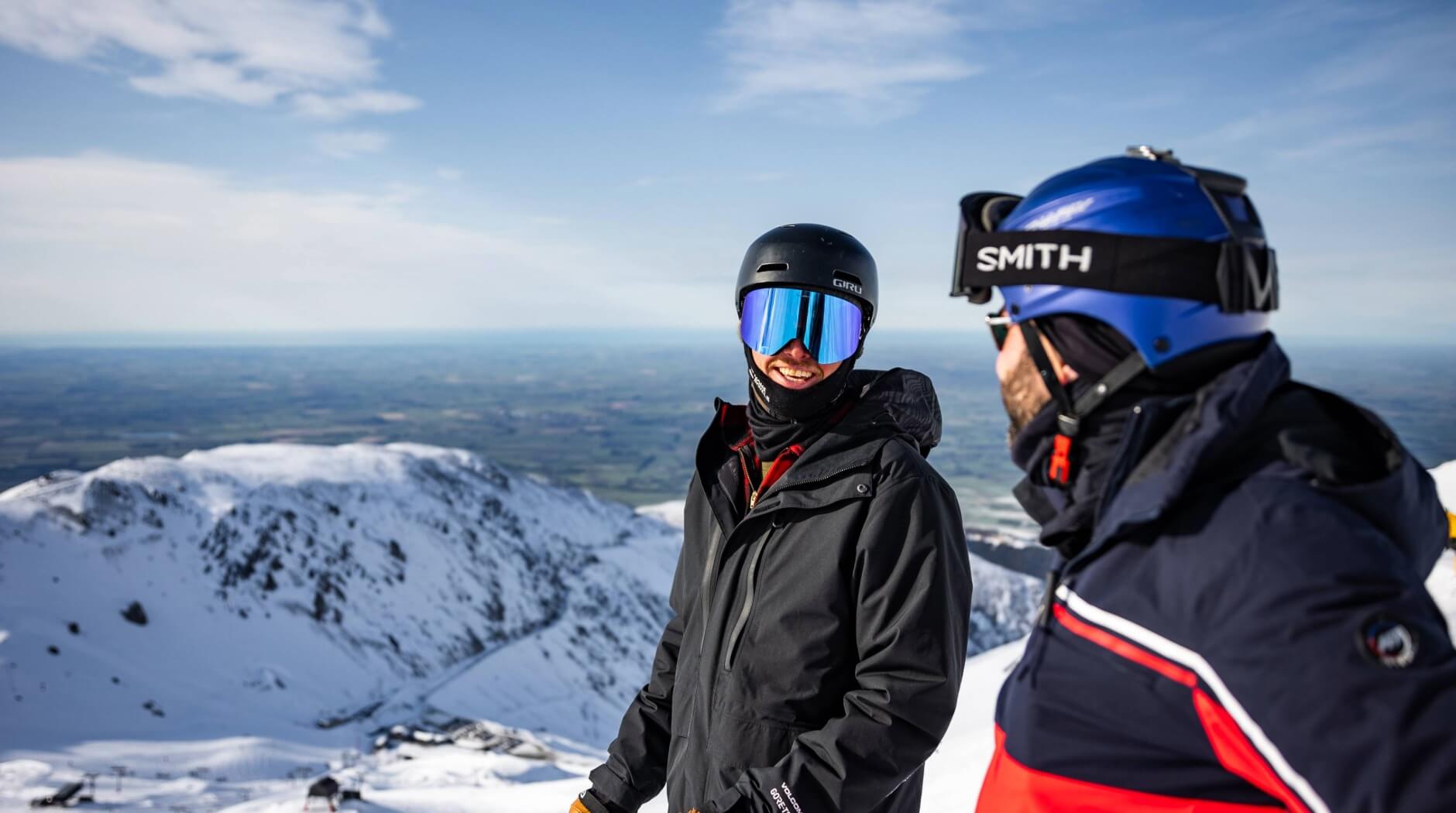 Two skiers in winter gear and helmets smiling on a snowy mountain with a clear sky and distant landscape.