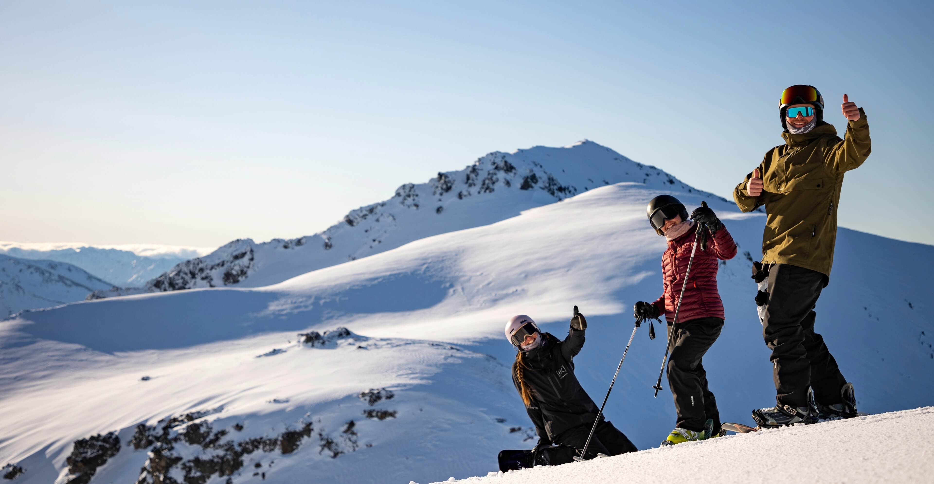 Three skiers on a snowy mountain slope giving thumbs up with snow-covered peaks in the background.