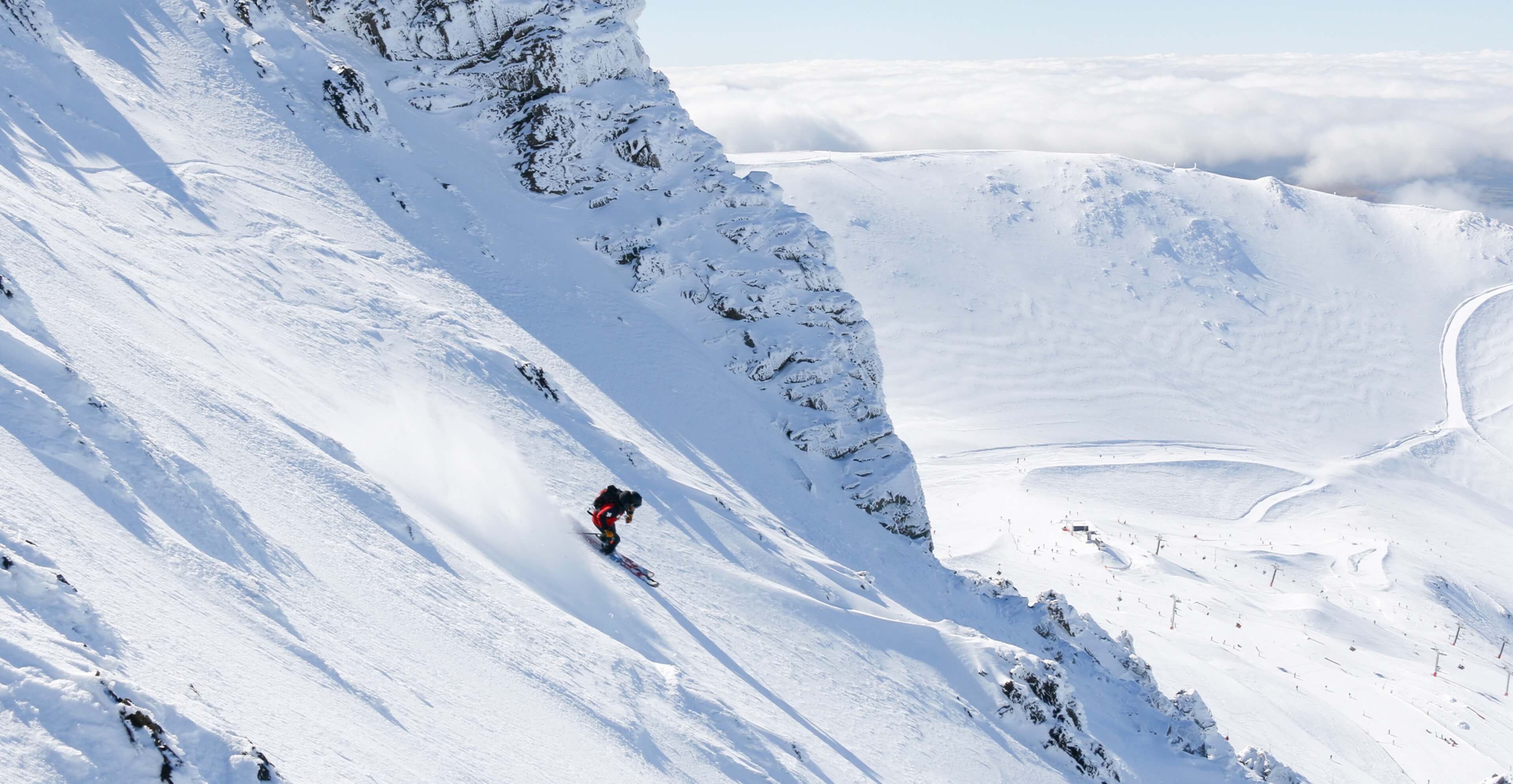 Skier descending steep snowy mountain slope with rocky outcrop and ski resort visible below.