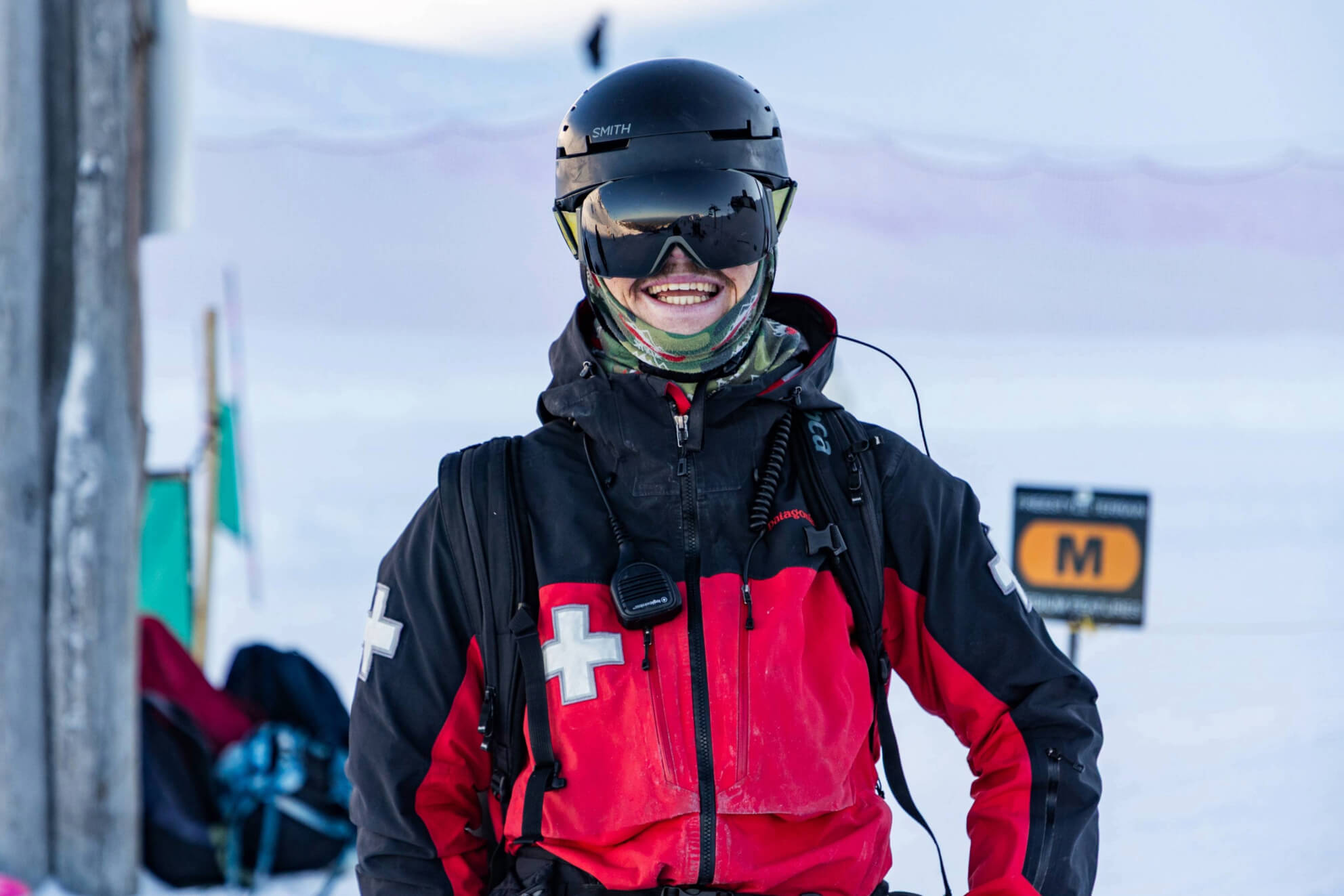 Smiling ski patrol member wearing a red and black jacket with a white cross, black helmet, and reflective goggles in a snowy mountain setting.