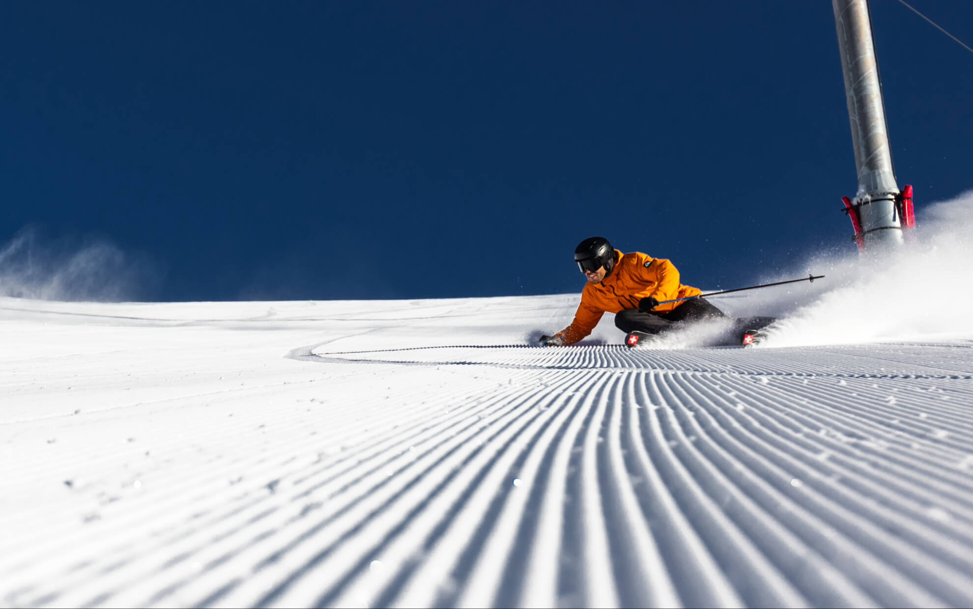 Skier in an orange jacket carving fresh tracks on groomed snow under a clear blue sky.