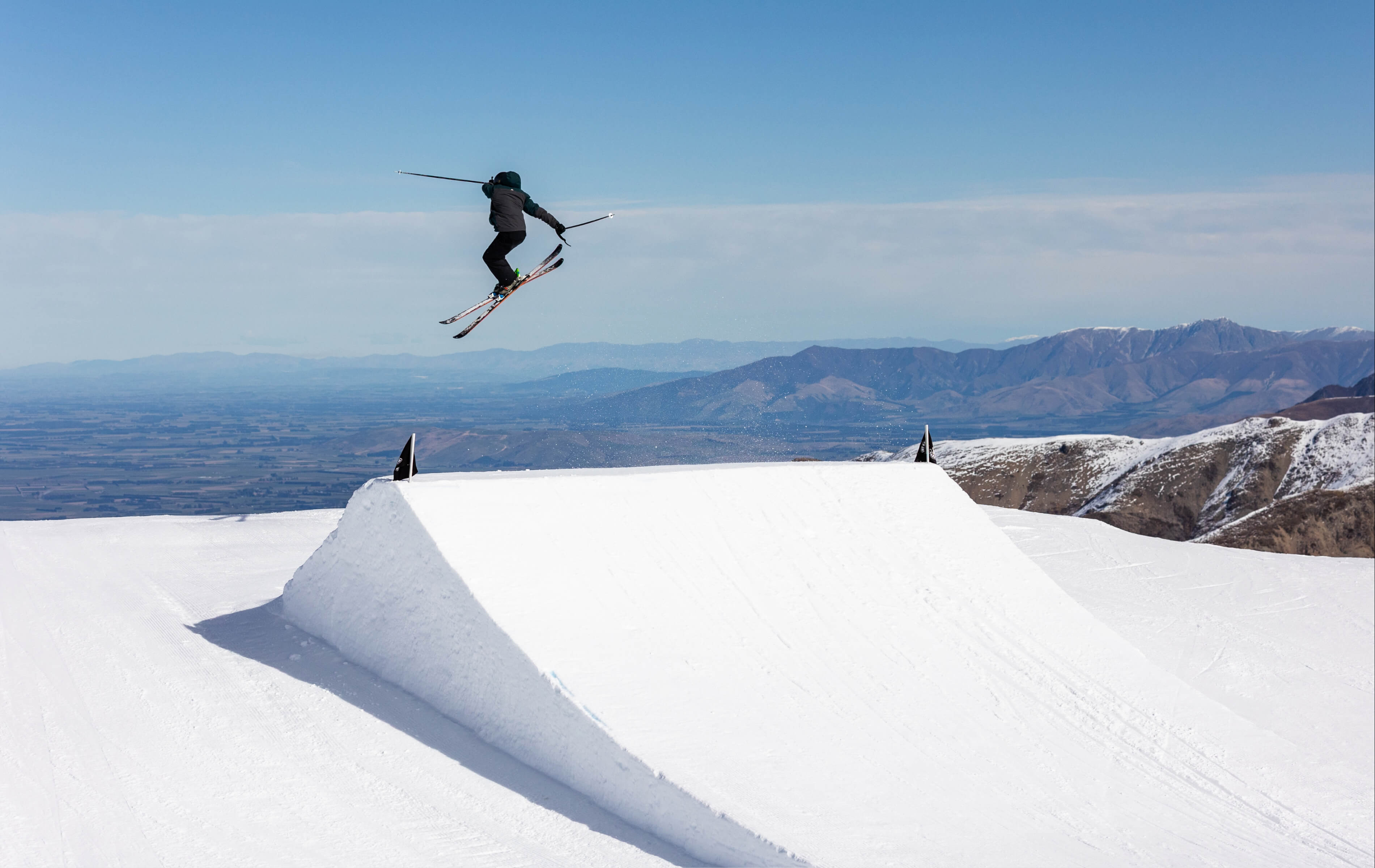 Skier performing a jump trick in the air over a large snow ramp in a mountainous ski park under a clear blue sky.