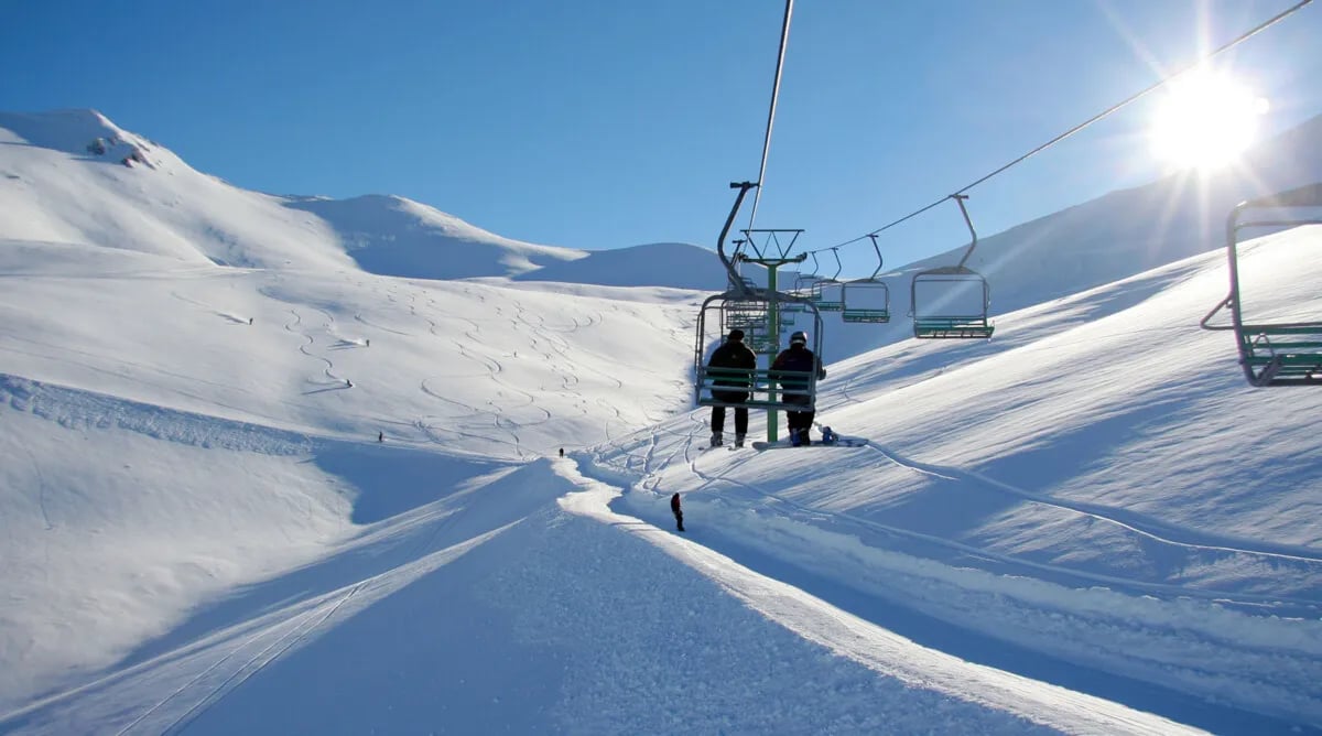 Ski chairlift carrying two people over snow-covered mountains under a clear blue sky with bright sunlight.