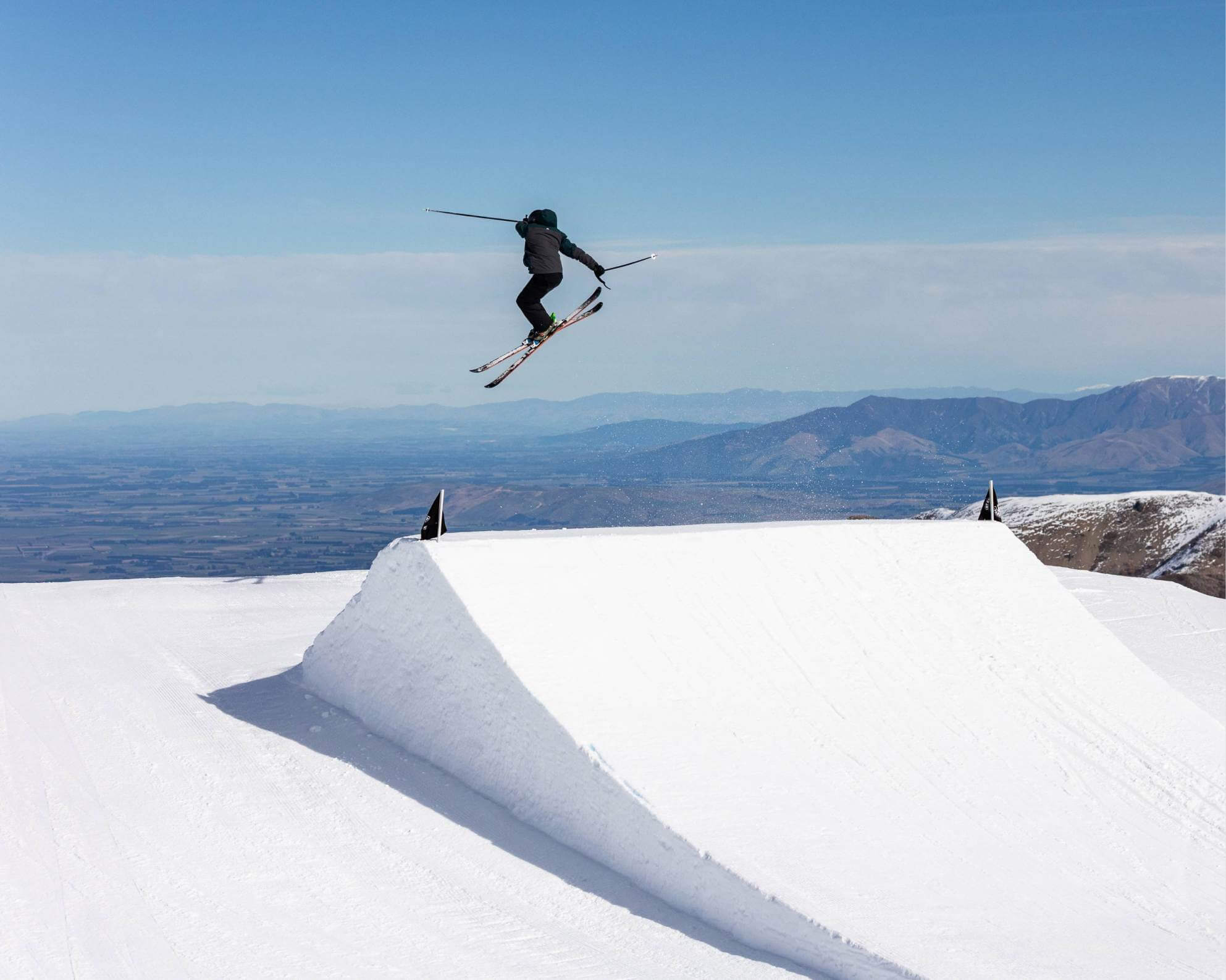 Skier performing a jump trick in mid-air over a snow ramp with mountain landscape in the background.