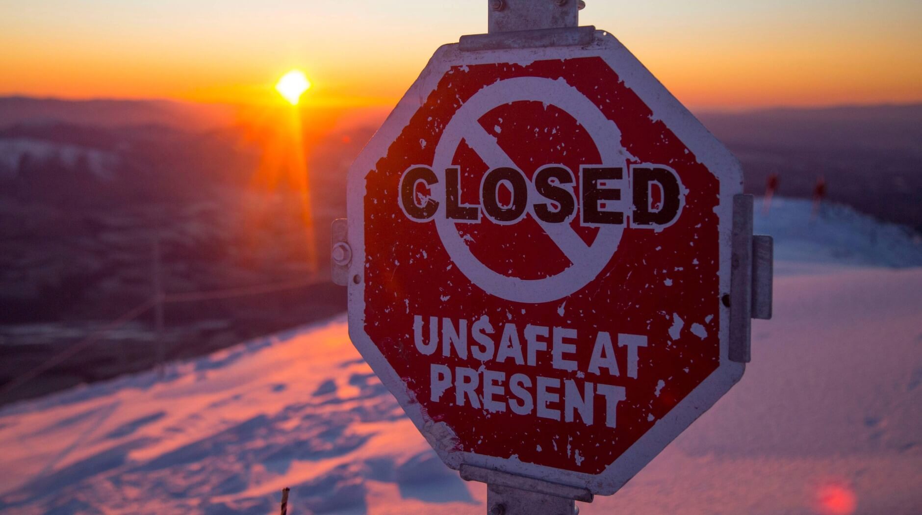 Weathered red octagonal sign reading 'CLOSED UNSAFE AT PRESENT' on a snowy mountain at sunset.