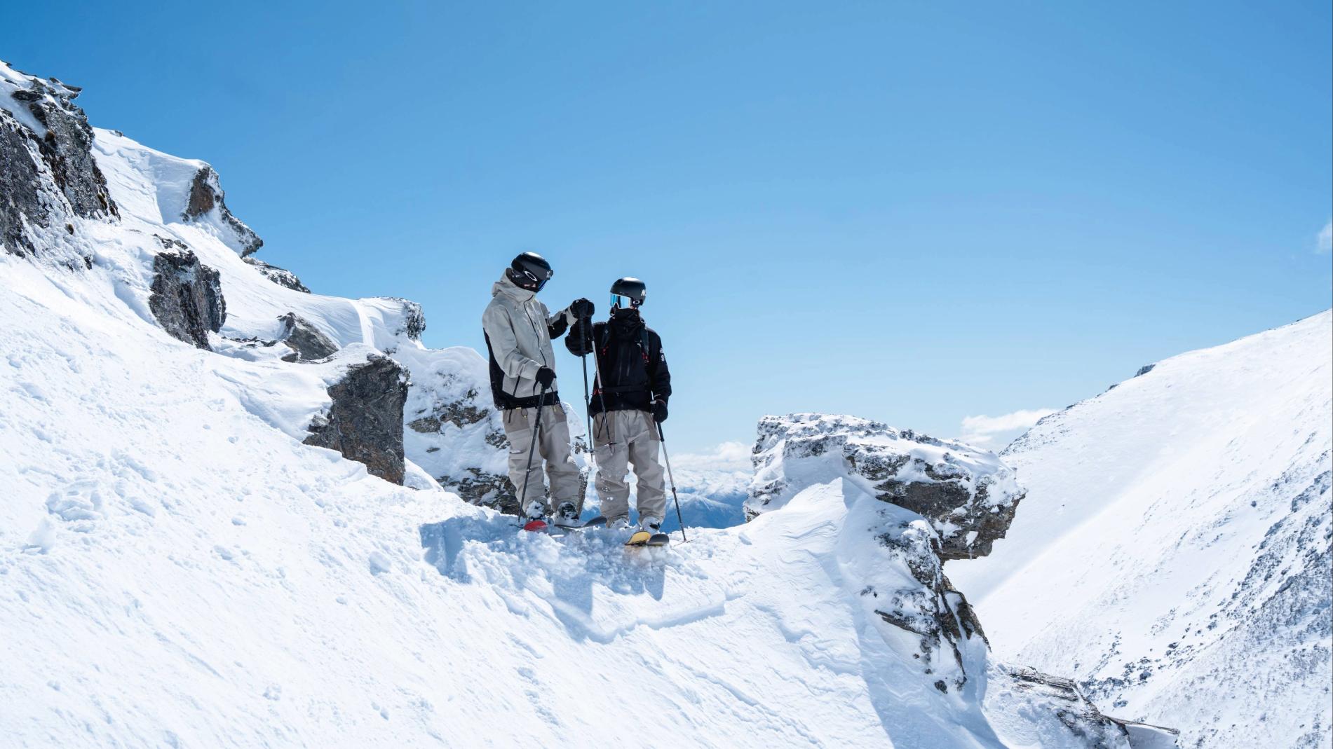 Two skiers in helmets and ski gear standing on a snowy mountain ridge with blue sky background.